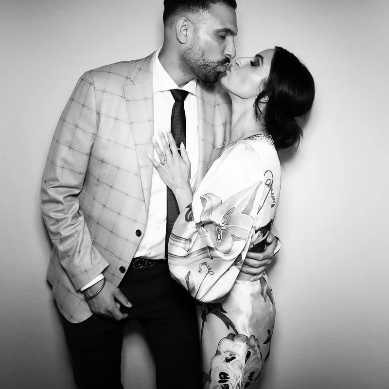A man and woman sharing a kiss, with the man wearing a checked suit and the woman in a floral dress, against a plain background in black and white.