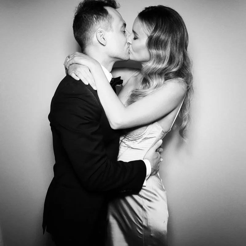 A black and white photo of a couple kissing, with the man wearing a tuxedo and the woman in a satin dress.