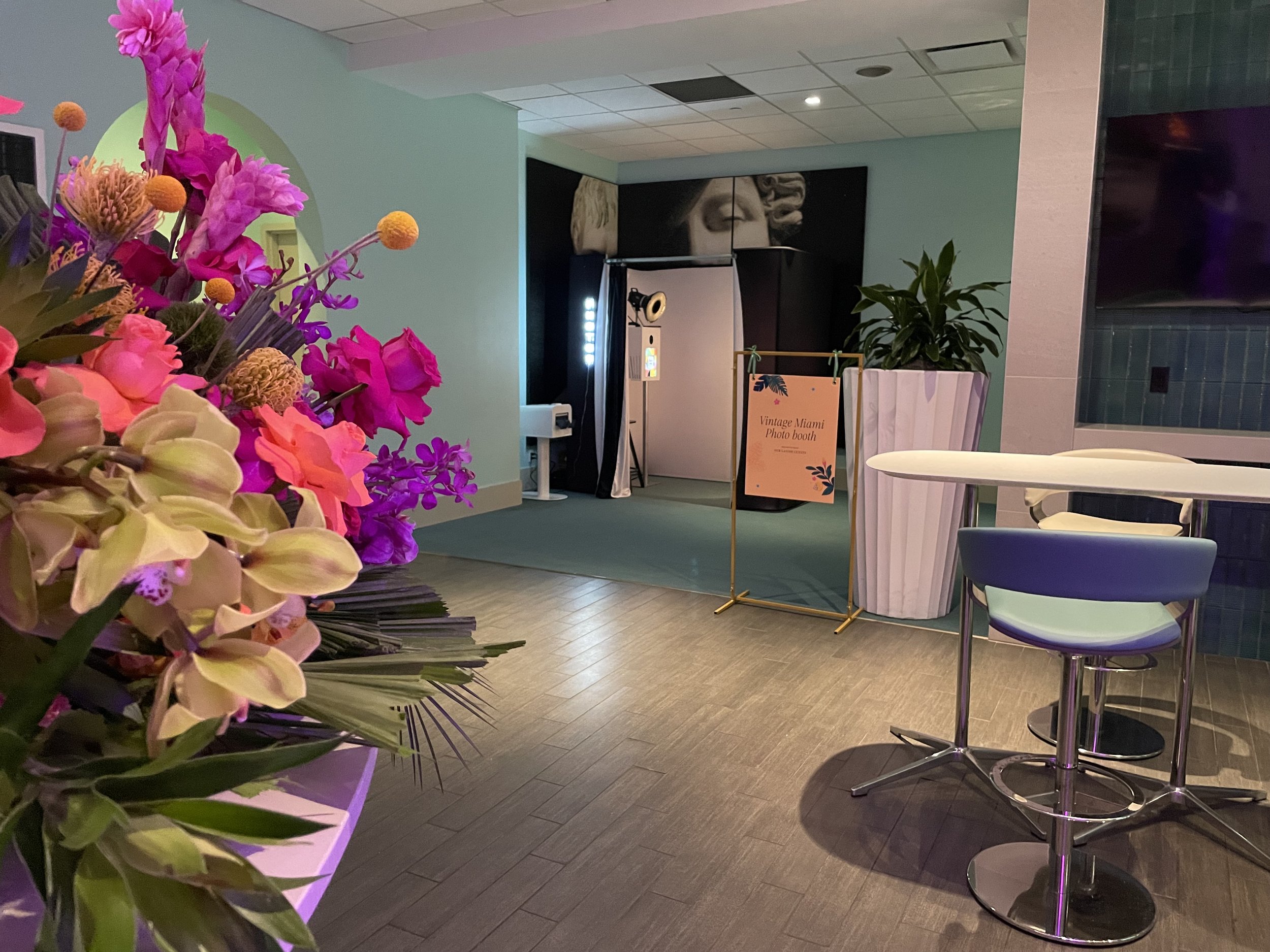 Indoor photo of a tropical-themed photo booth station with a sign that reads "Vintage Miami Photo booth." There is a large potted plant beside the sign and a framed picture of a classical sculpture in the background. A round table with a light-colore