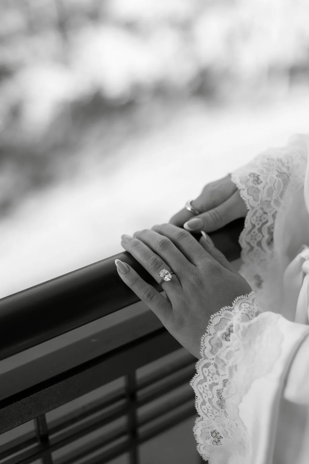 Close-up of a bride's hand with a wedding ring touching a railing, dressed in lace sleeves, in black and white.