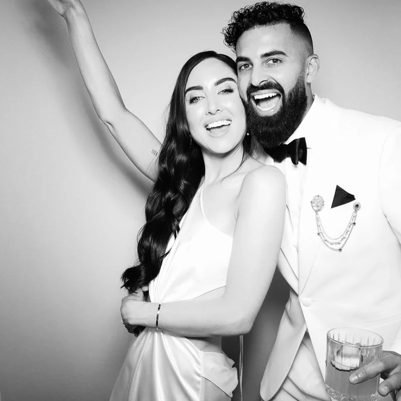 A black and white photo of a happy couple posing together. The woman has long dark hair and is wearing a slip dress. The man has a beard, curly hair, and is wearing a tuxedo with a bow tie, holding a glass of drink. They are smiling and the woman is 