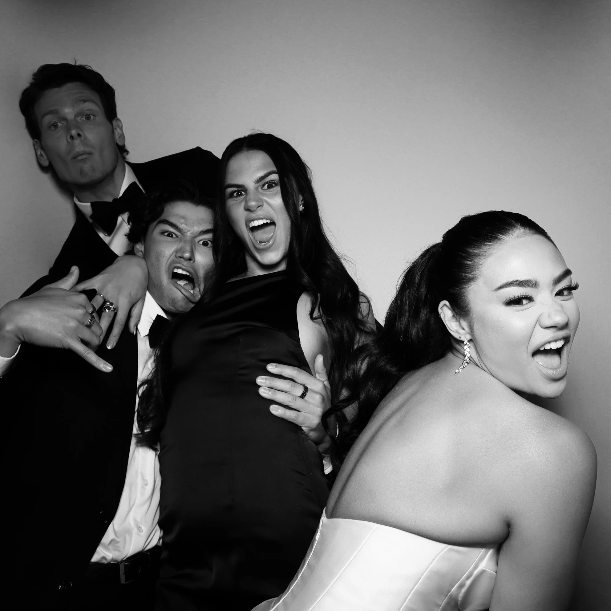 Black and white photo of four young adults dressed in formal attire, expressing playful and energetic moods, with varied facial expressions and poses.