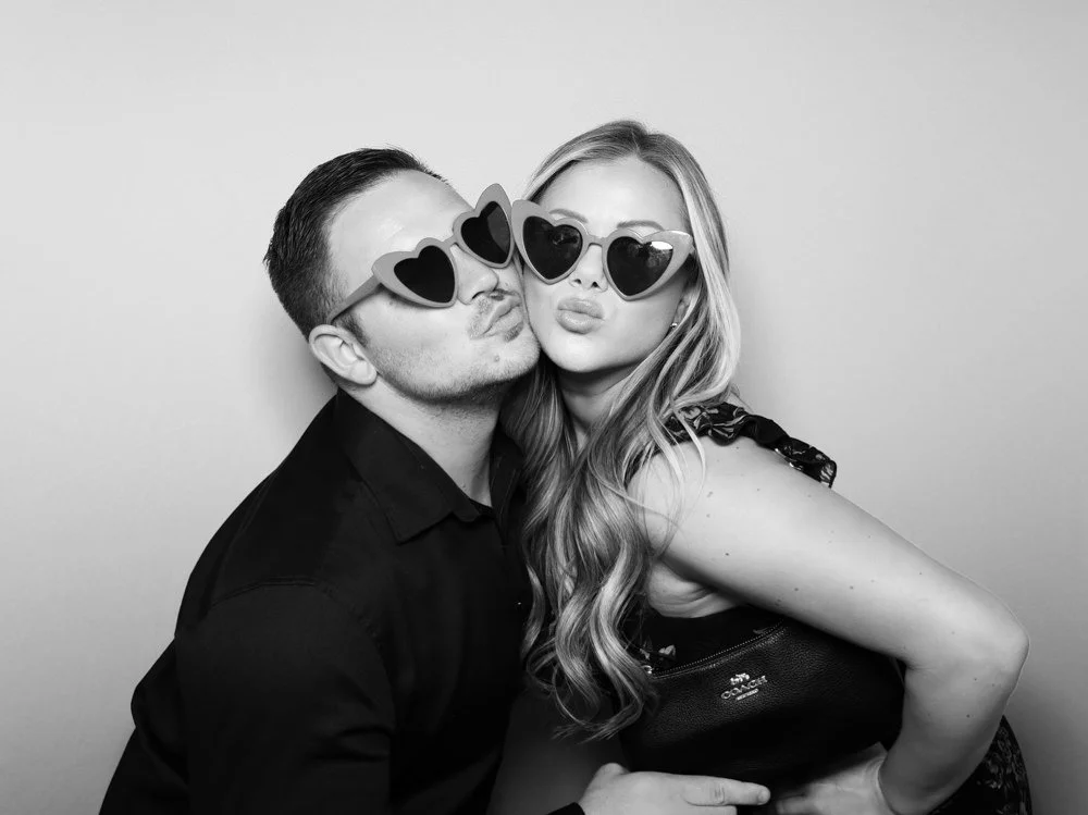 A man and a woman wearing heart-shaped sunglasses, kissing and posing for a photo, in black and white.