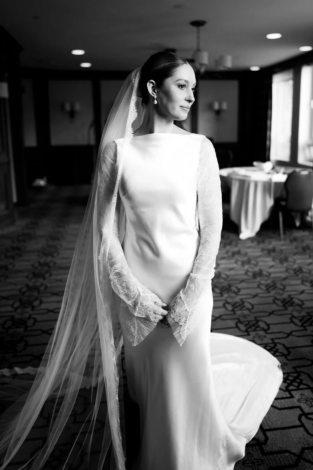 A bride in a wedding dress with lace long sleeves, wearing a veil and pearl earrings, standing in a hotel or banquet room with tables and chairs in the background.