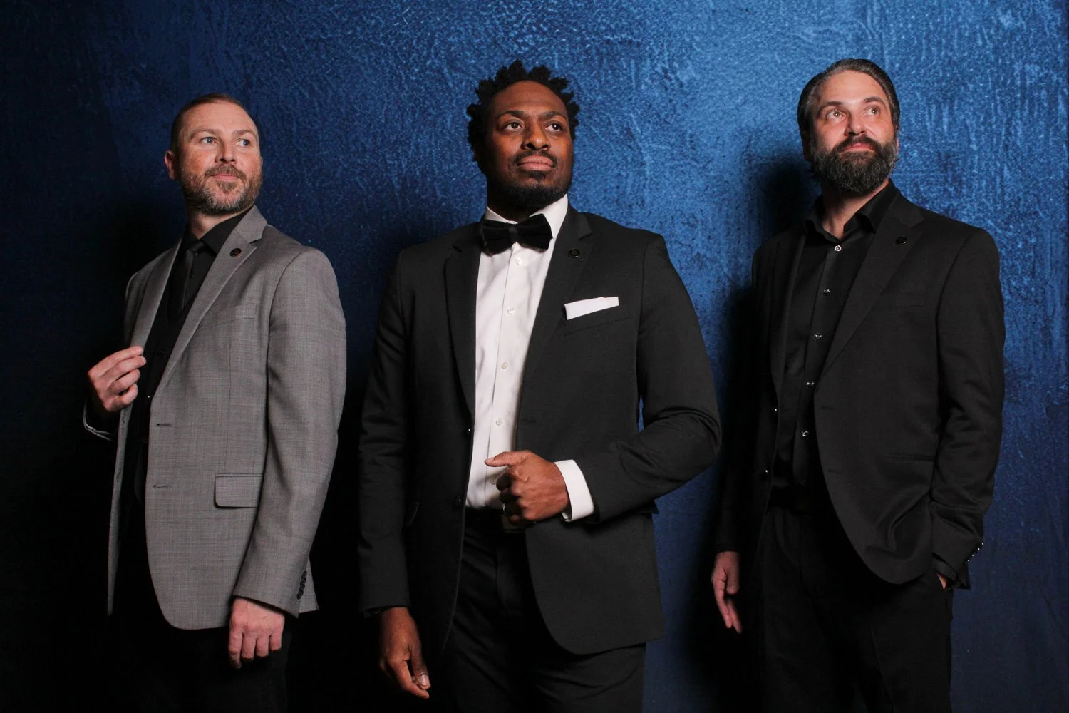 Three men in formal attire standing against a blue textured wall. The man on the left is wearing a light gray checked suit and black shirt. The man in the center is wearing a black tuxedo with a bow tie and white shirt. The man on the right is wearin