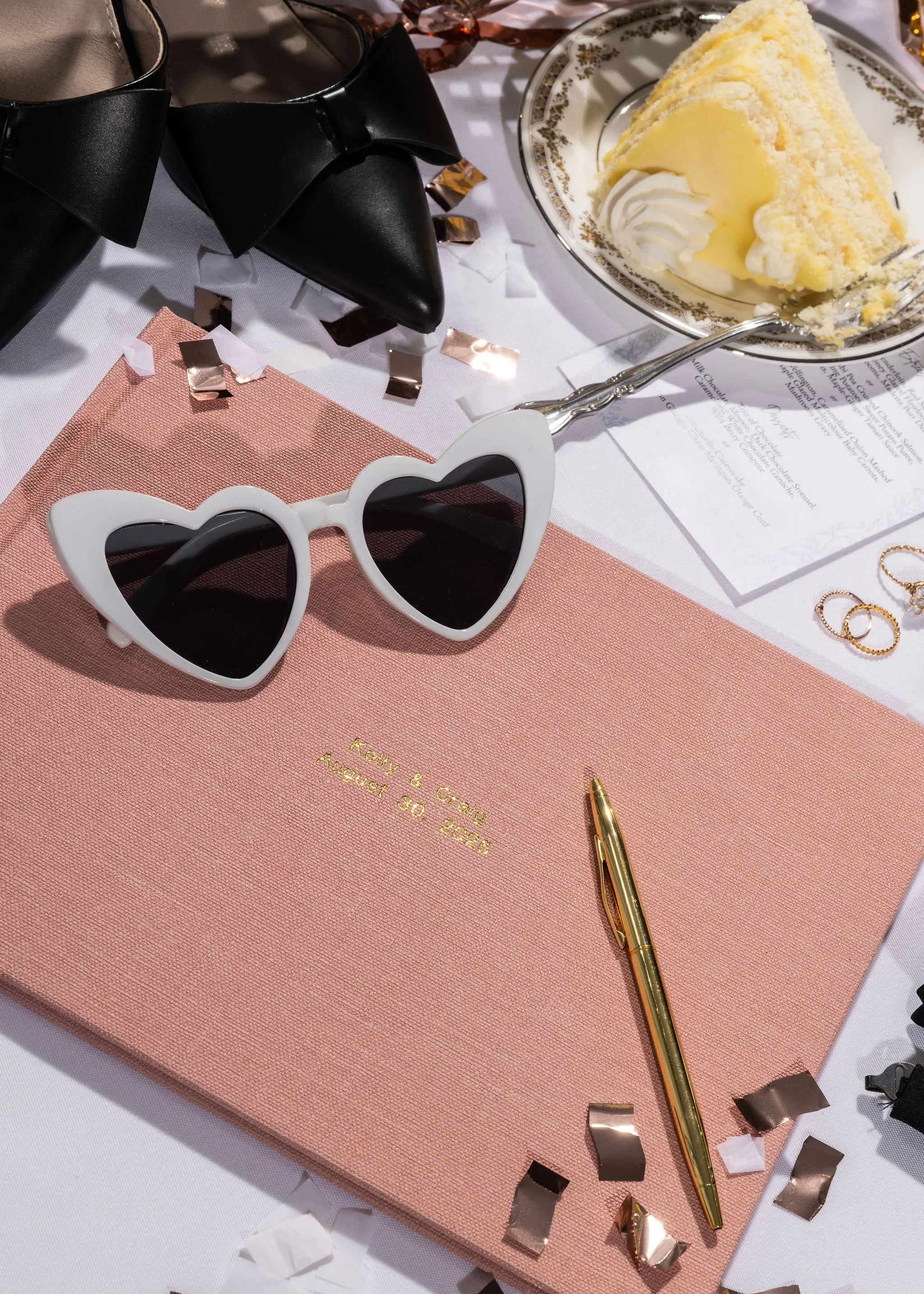 A pink wedding guest book with gold embossed text, white heart-shaped sunglasses, a gold pen, a slice of birthday cake with whipped cream and icing on a decorated plate, a black bow tie, gold rings, and scattered confetti on a table with a white tabl