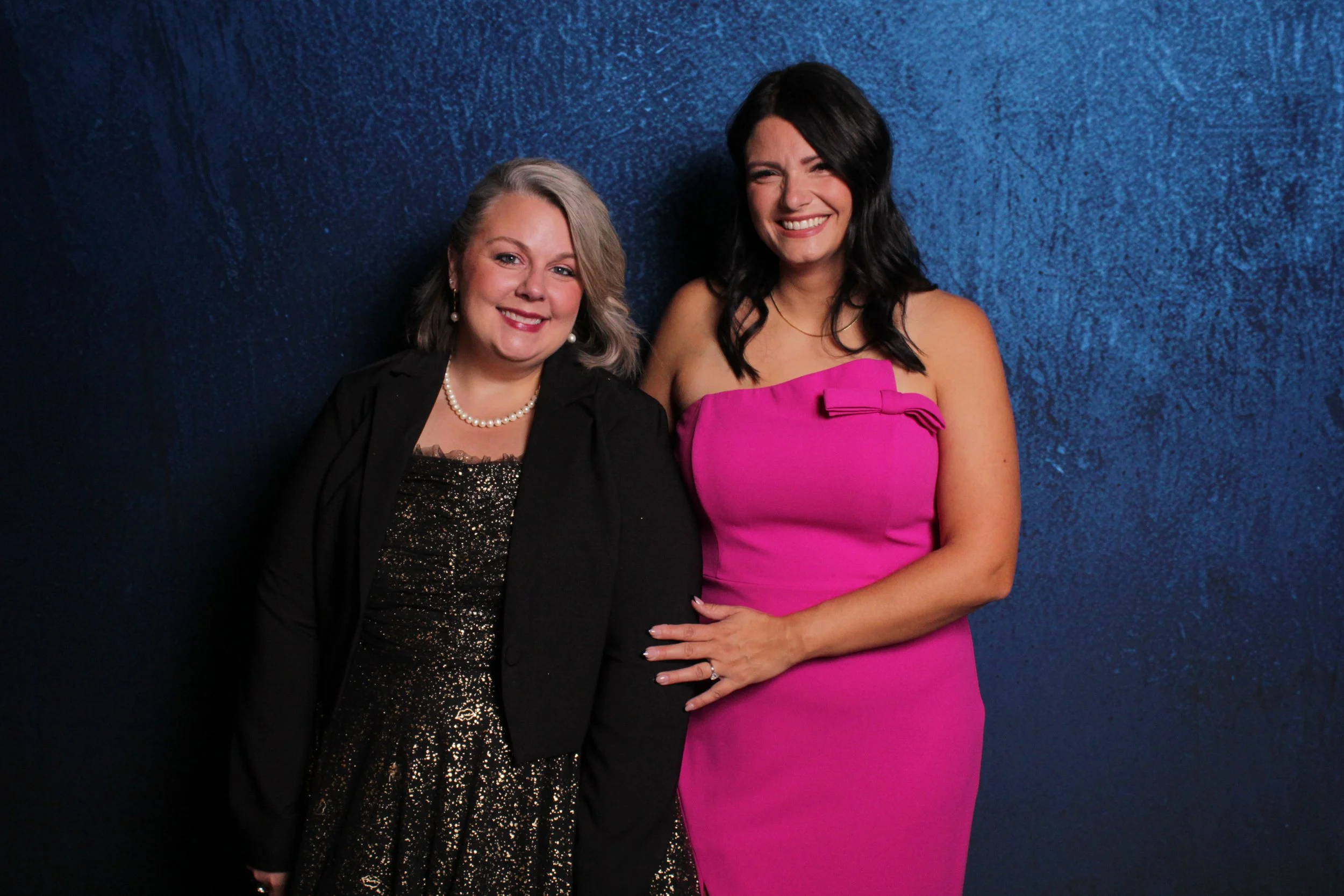 Two women smiling in formal attire, one wearing a black and gold dress with a blazer and pearl necklace, the other wearing a bright pink strapless dress with a bow detail, standing against a textured blue background.