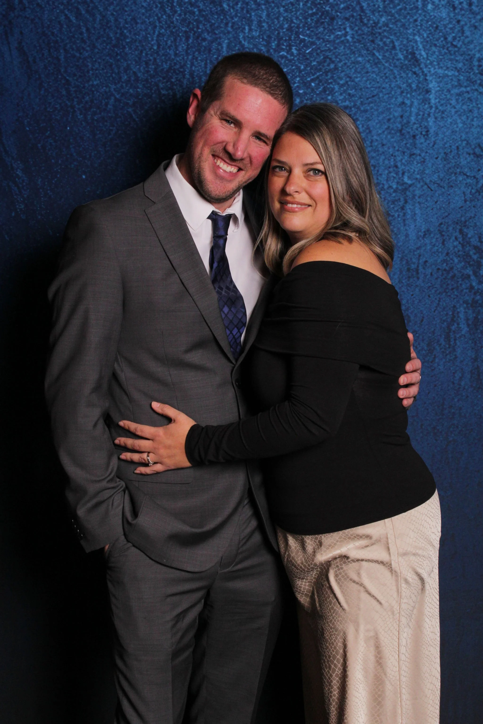 A man in a gray suit and a woman in a black top and beige pants pose together against a blue textured background