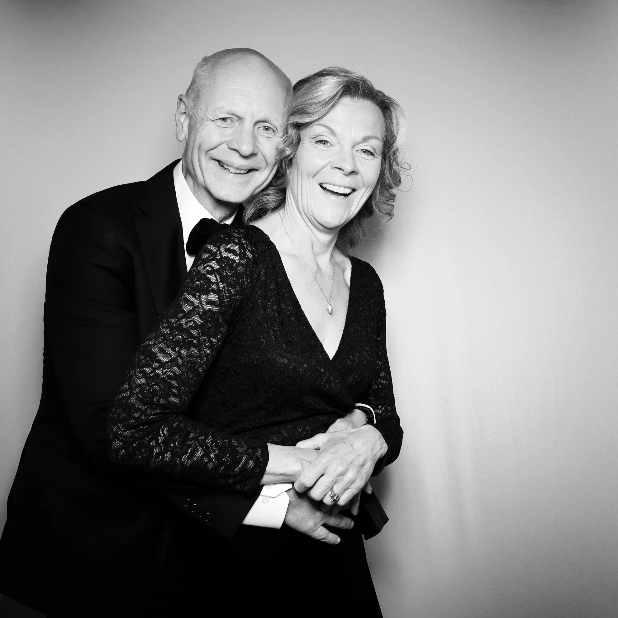 Black and white photo of a smiling older man in a tuxedo with bow tie, embracing a smiling woman in a black lace dress, standing against a plain wall.