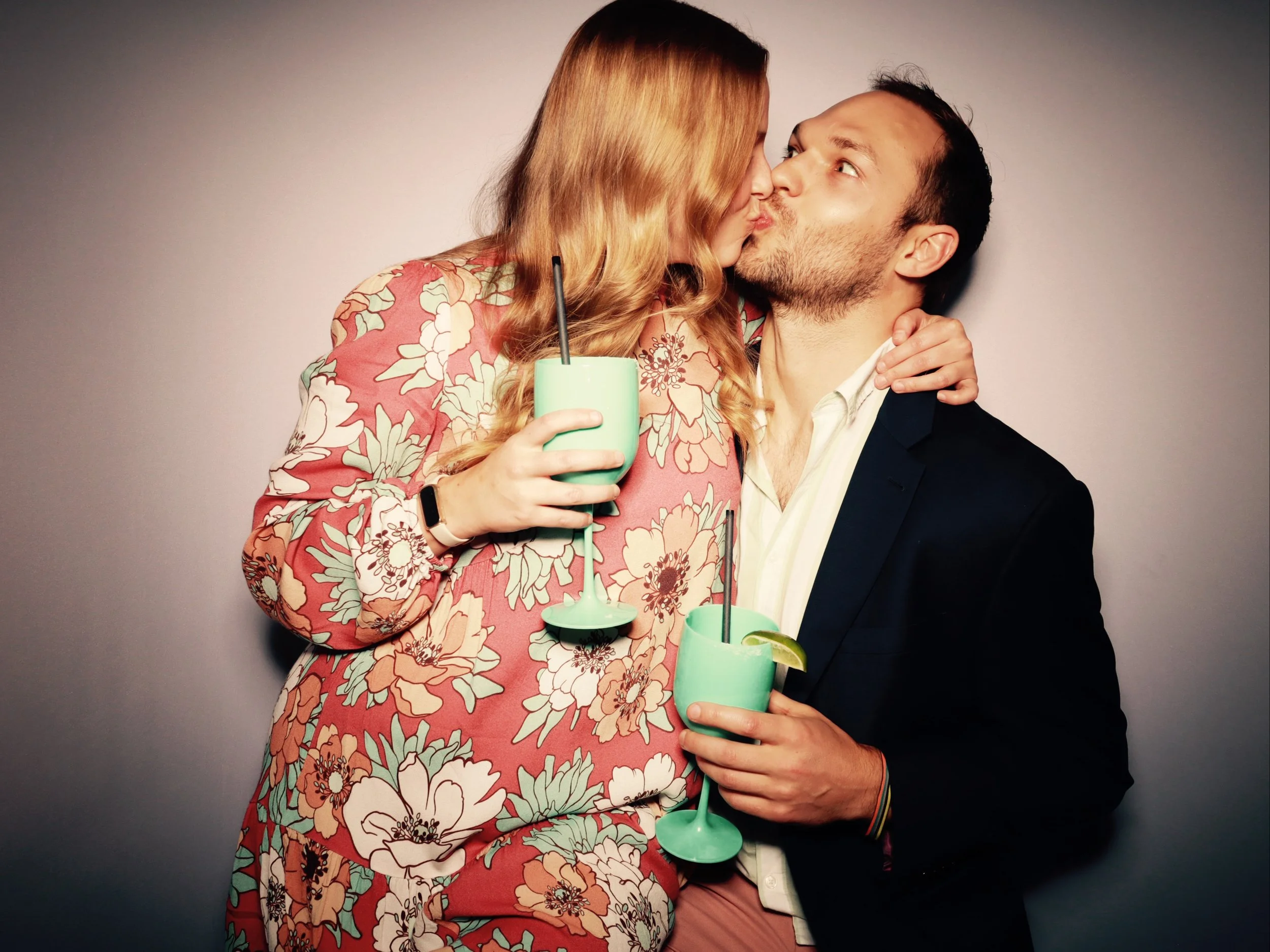 A man and woman share a kiss while holding green cocktails with lime wedges and straws. The woman has red hair and wears a floral dress, and the man has dark hair and wears a suit jacket over a white shirt.