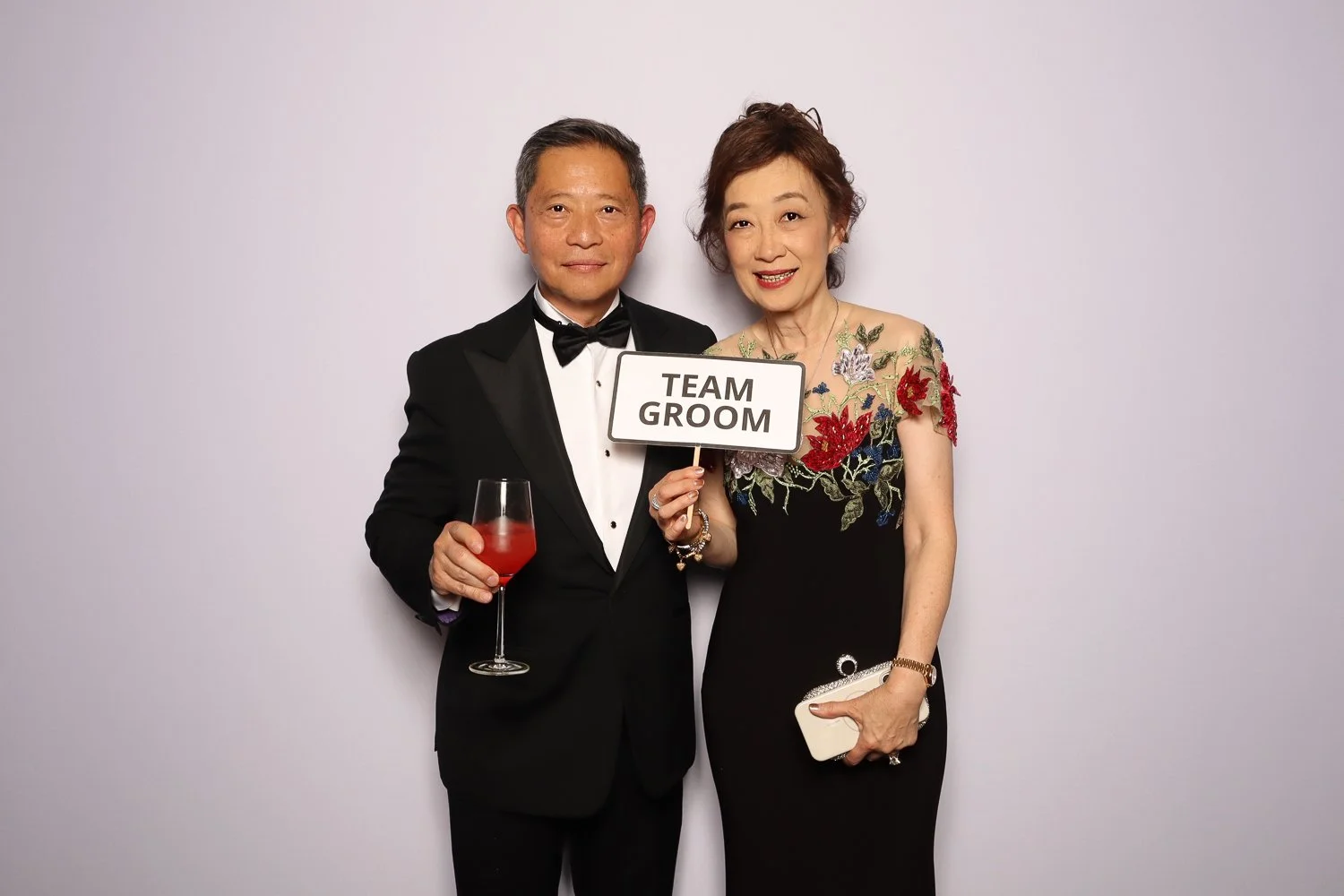 A happy middle-aged couple dressed in formal attire, holding a sign that says 'TEAM GROOM' at a wedding or event.