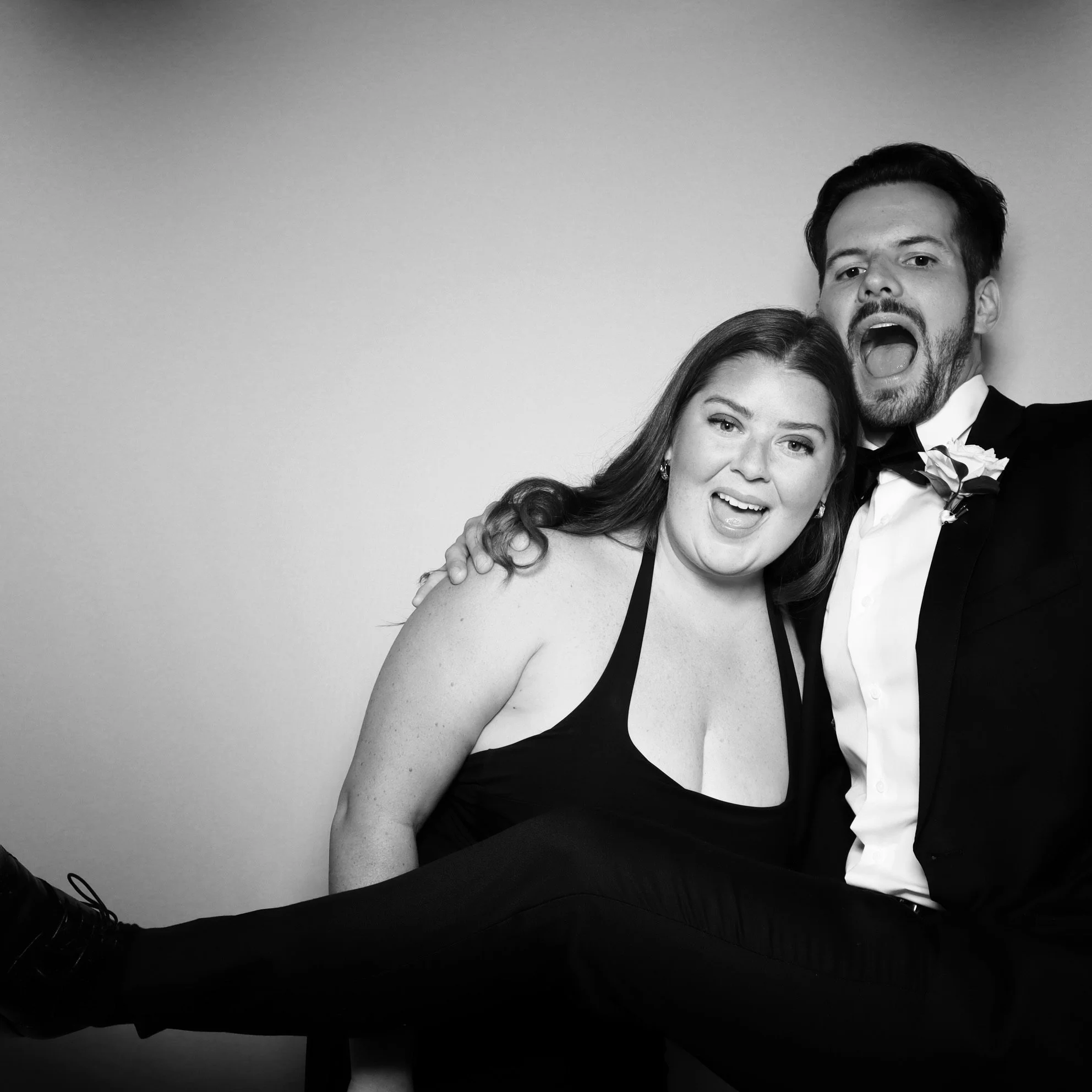 Black and white photo of a couple at a formal event, with the woman sitting on the man's lap. Both are smiling and making playful faces, dressed in tuxedo and evening dress.