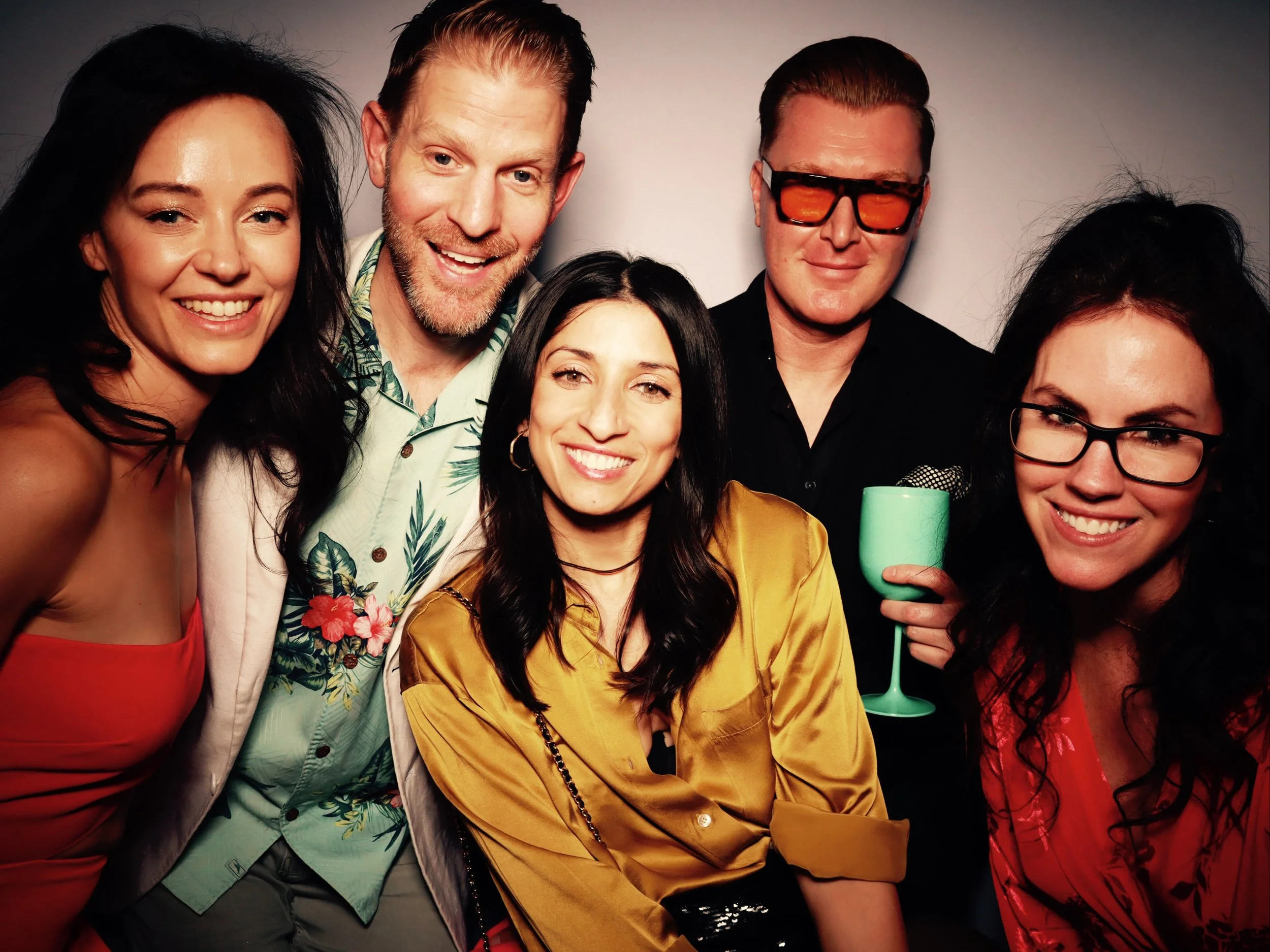 Group of five people smiling at a party, one person holding a microphone, in a relaxed setting.
