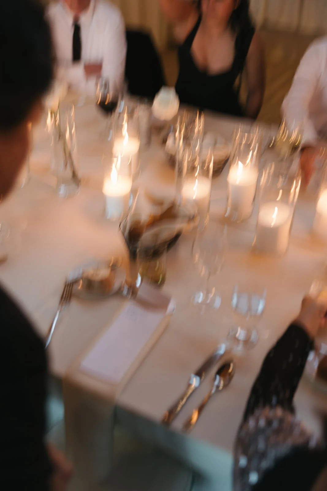 A blurry photo of a dinner table with lit candles, glasses, plates, silverware, and people seated around it, with some people dressed in formal attire.