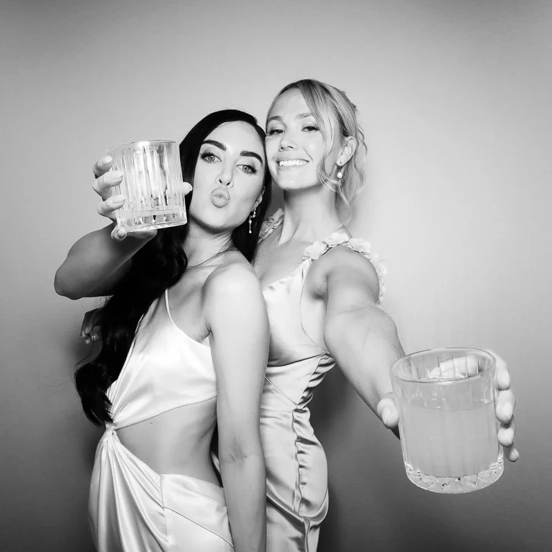 Two women in satin dresses taking a selfie, holding glasses of water, smiling and making kissy faces.