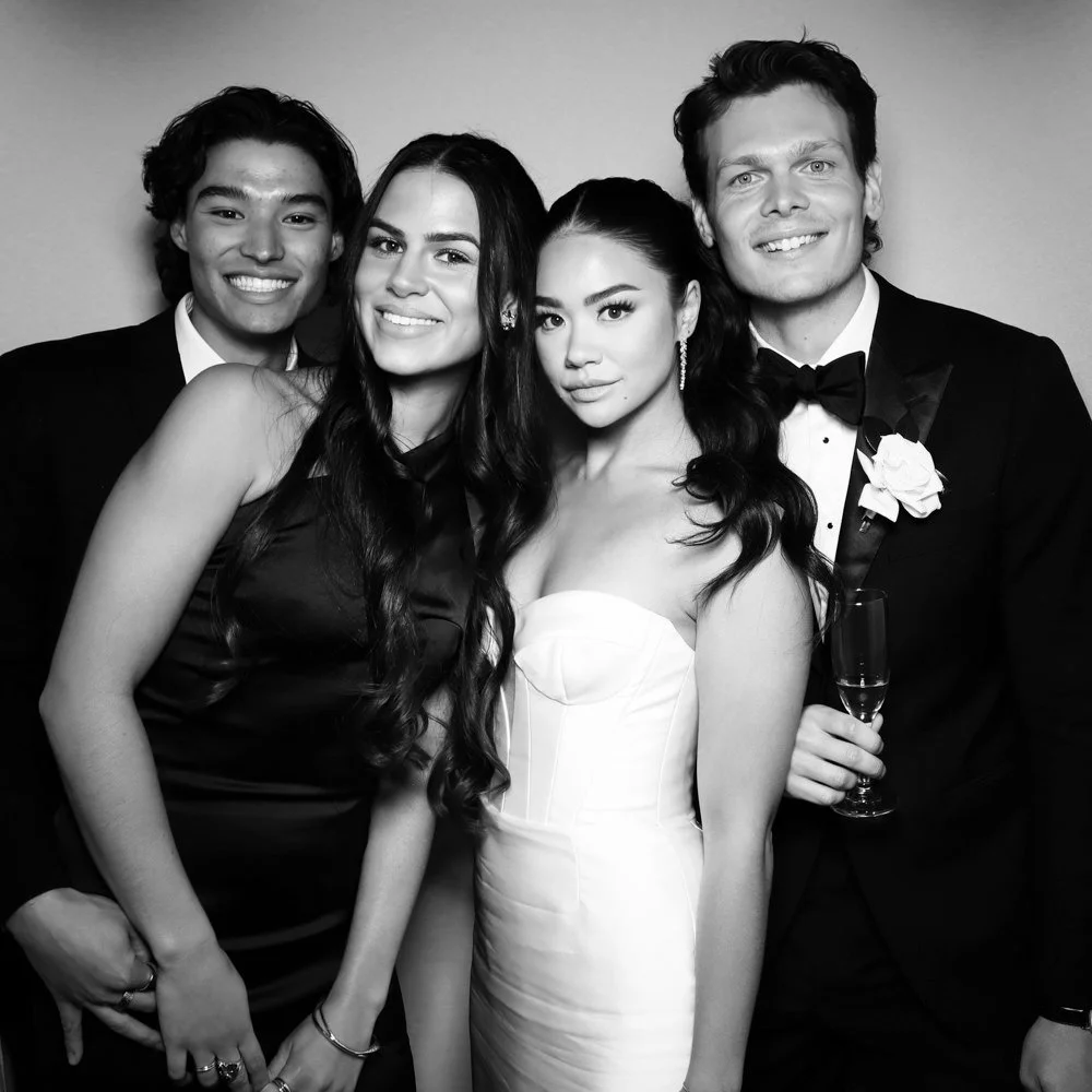 Black and white photo of four young adults dressed in formal attire at a celebration, with two men in tuxedos and two women in elegant dresses, standing close together and smiling.