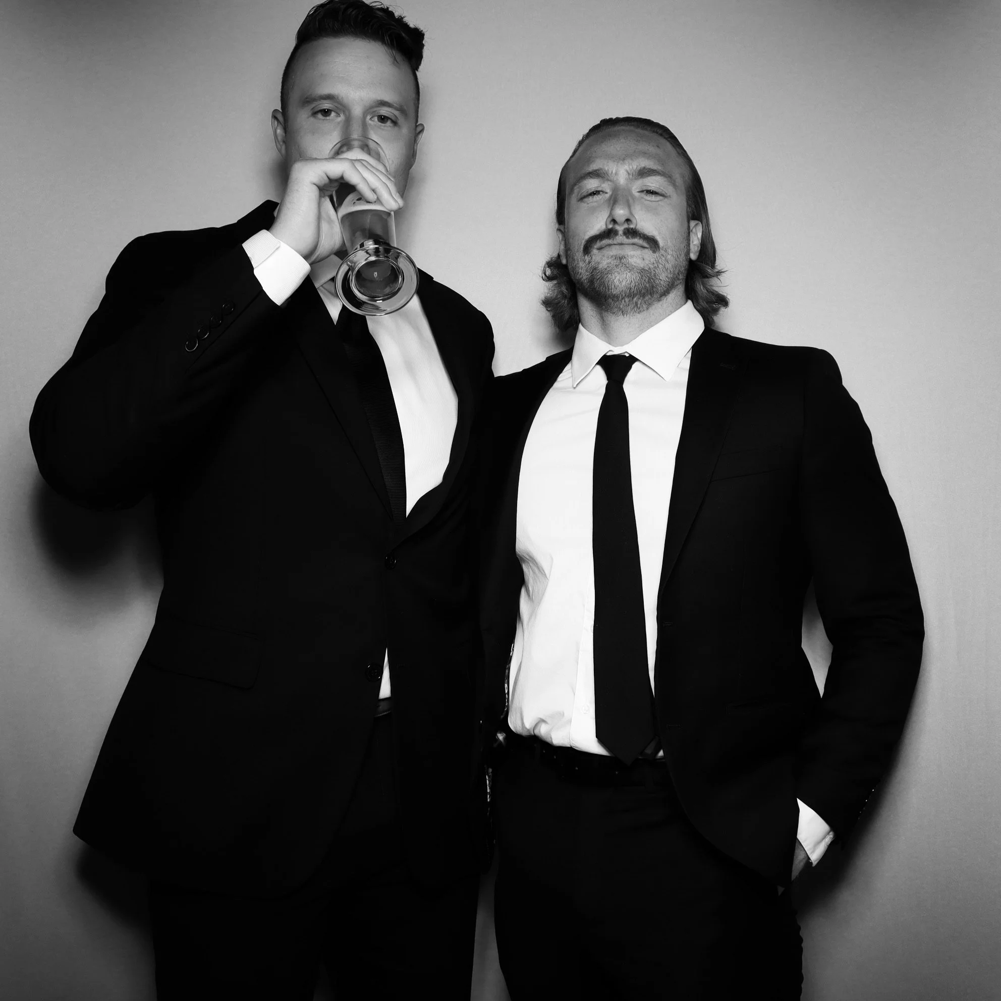 Two men in black suits and ties standing against a plain background, one drinking from a glass, both looking at the camera.