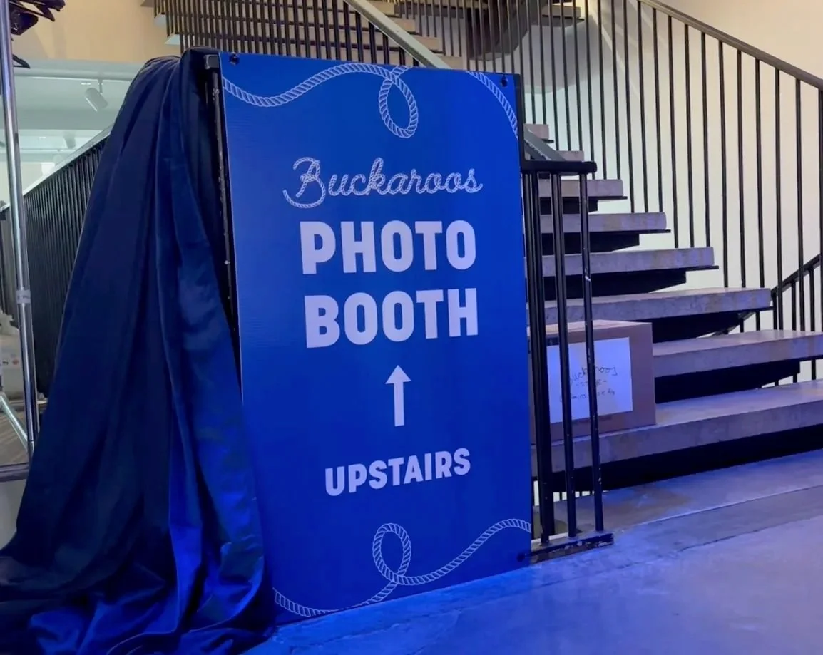 Blue sign indicating the location of the photo booth upstairs, with an arrow pointing upward. The sign is placed on the floor next to stairs with black railings.