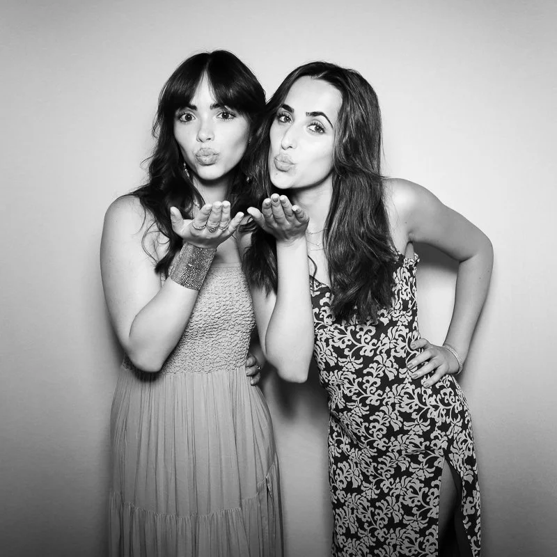 Two women blowing kisses toward the camera, standing close together against a plain background.