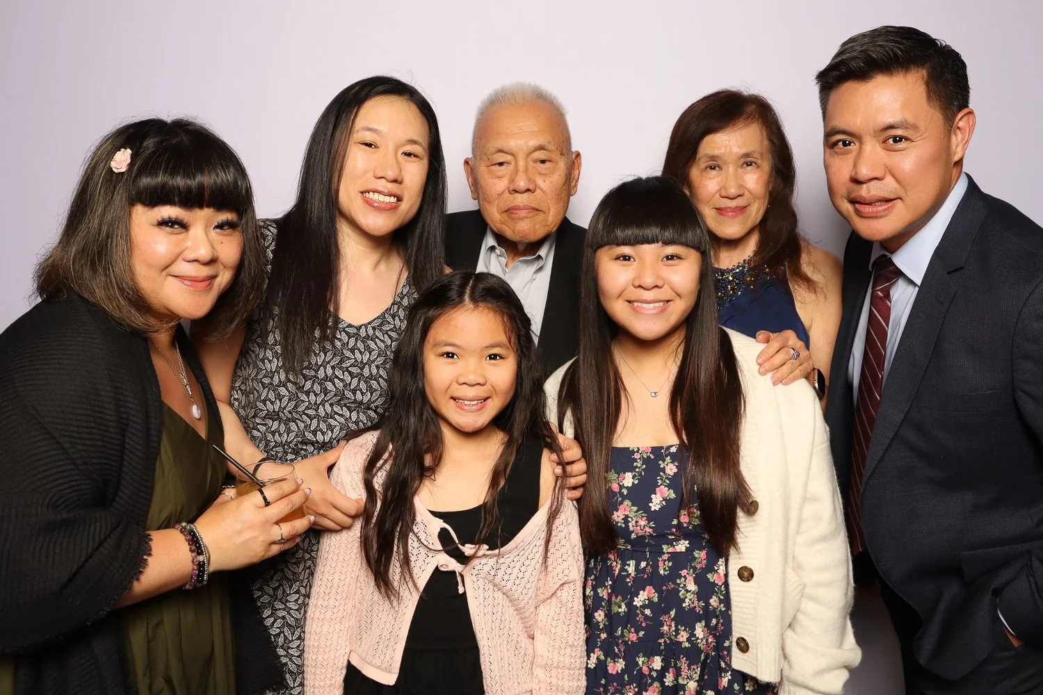 Family group portrait with seven members, including children and elderly, smiling and posing in front of a plain white background.