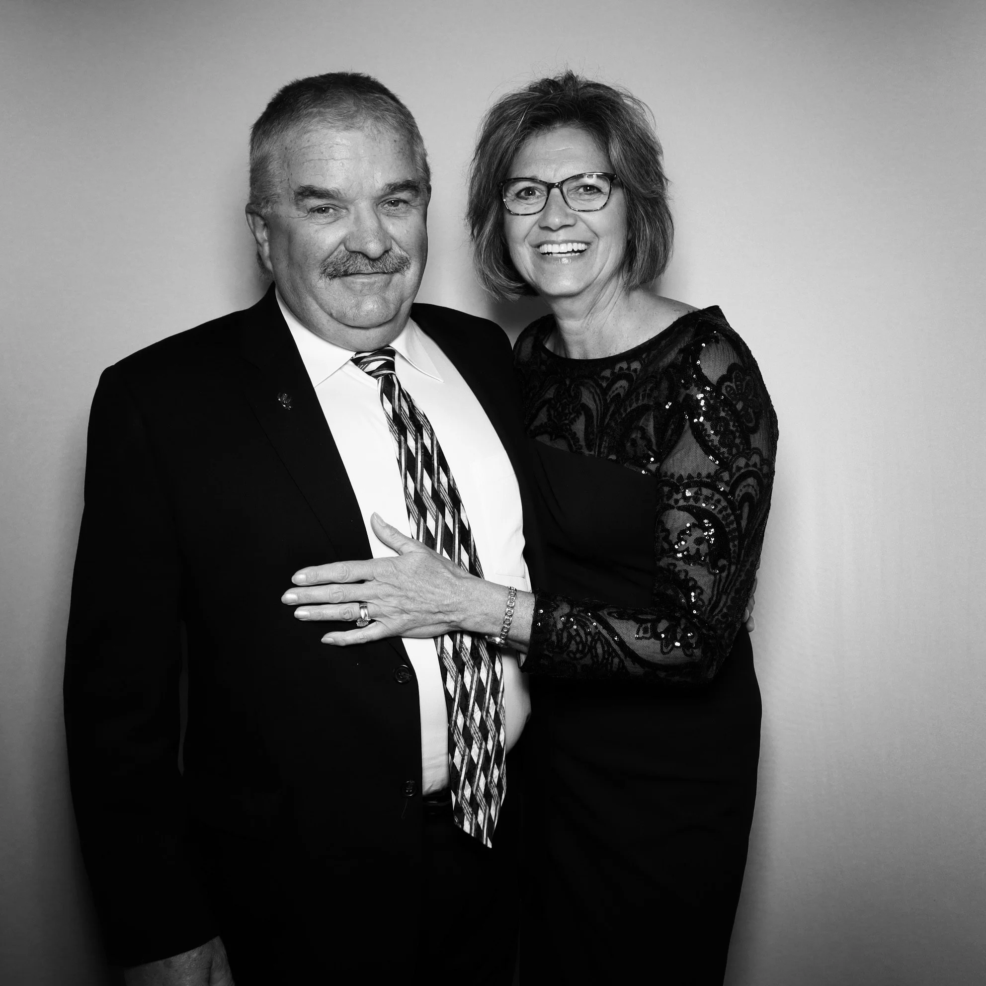 Black and white photo of a man and woman standing close together, smiling, with the woman holding the man's chest, against a plain background.
