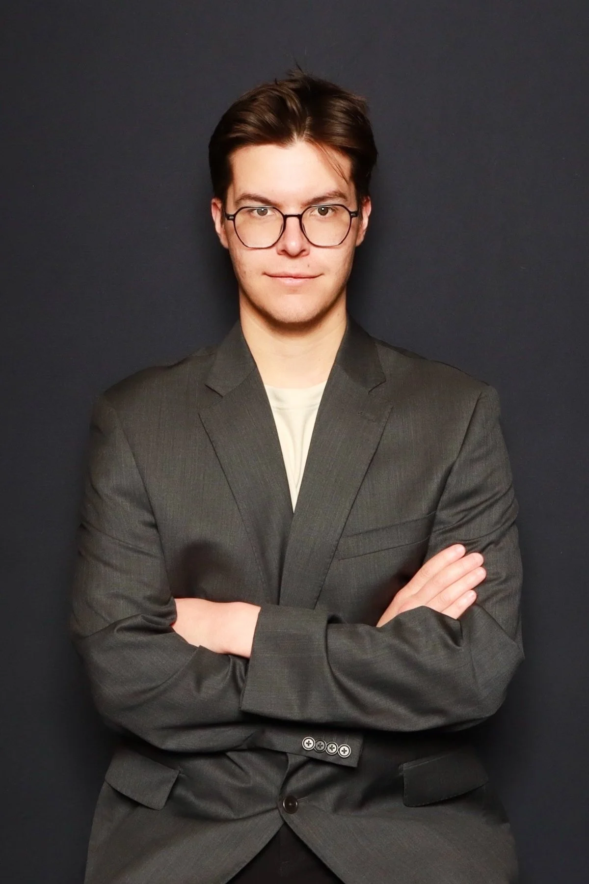 A young man with brown hair, wearing glasses, a dark gray suit, and a light-colored shirt, standing with arms crossed against a dark background.