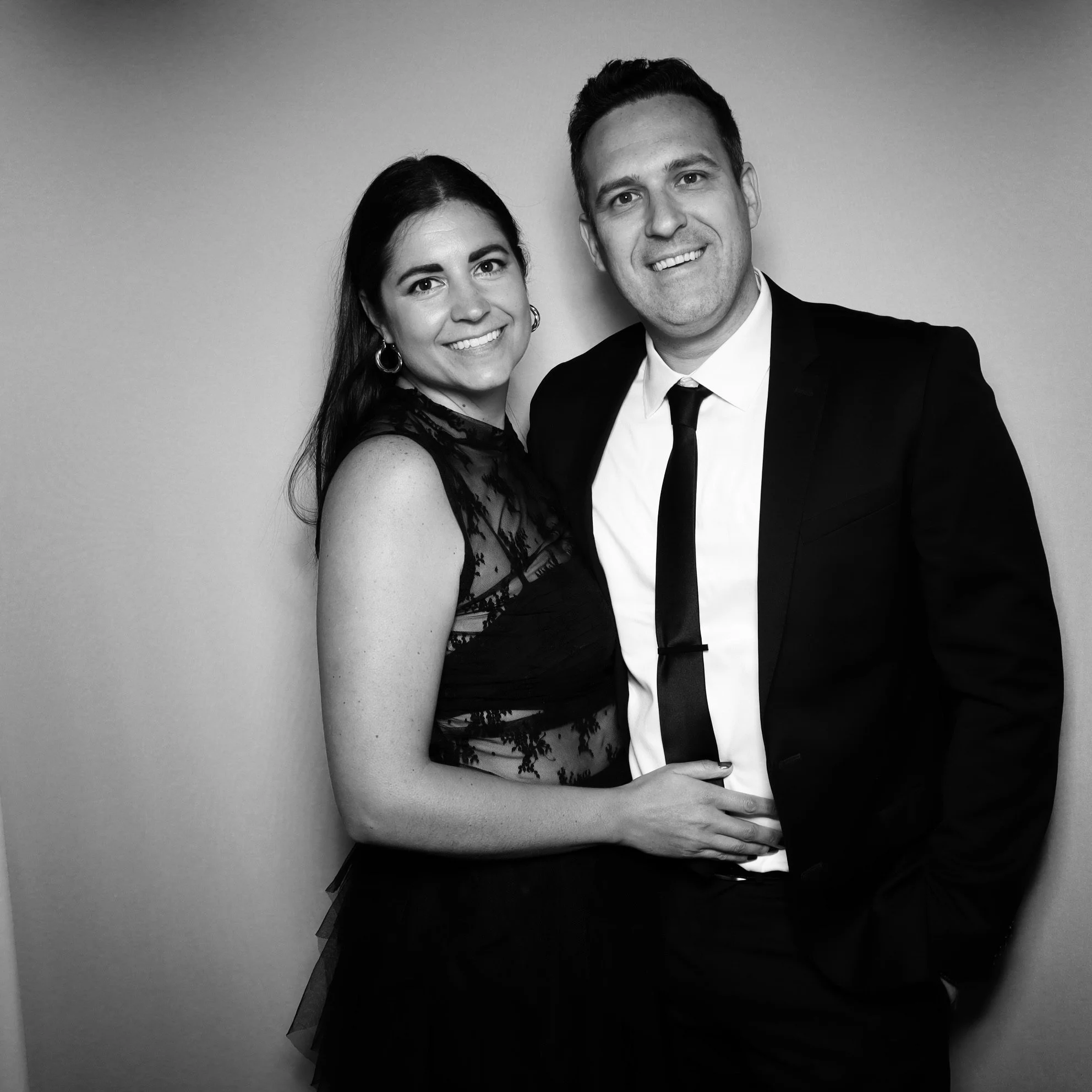 A black-and-white photo of a smiling couple dressed in formal attire, standing closely together against a plain background.