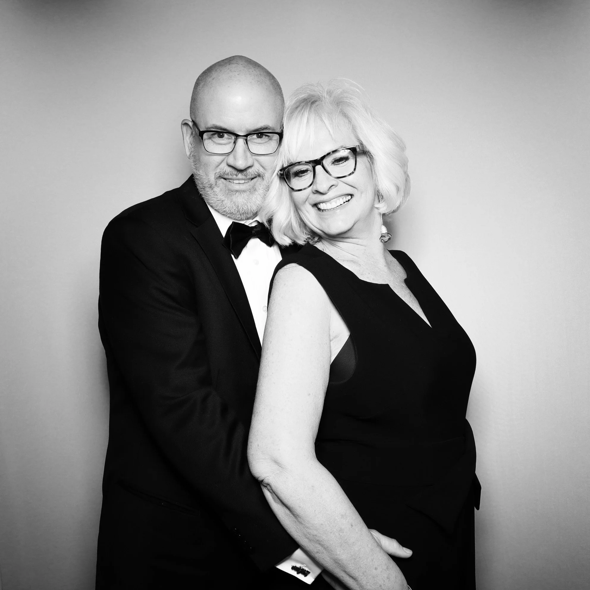 A black-and-white photograph of a smiling older woman and a man dressed in a tuxedo, both wearing glasses, embracing each other against a plain background.
