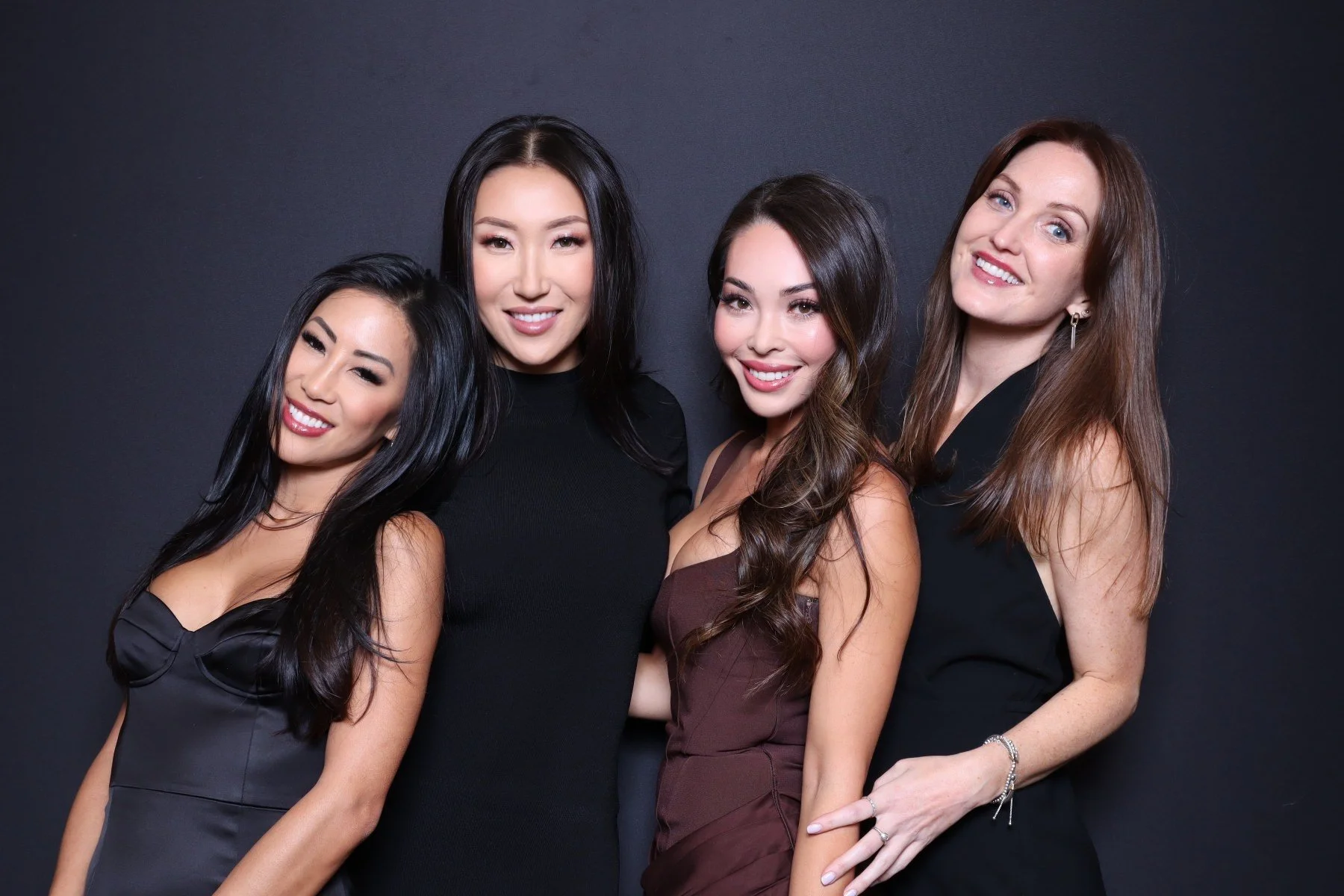 Four women dressed in black and brown dresses standing against a dark background, smiling at the camera.