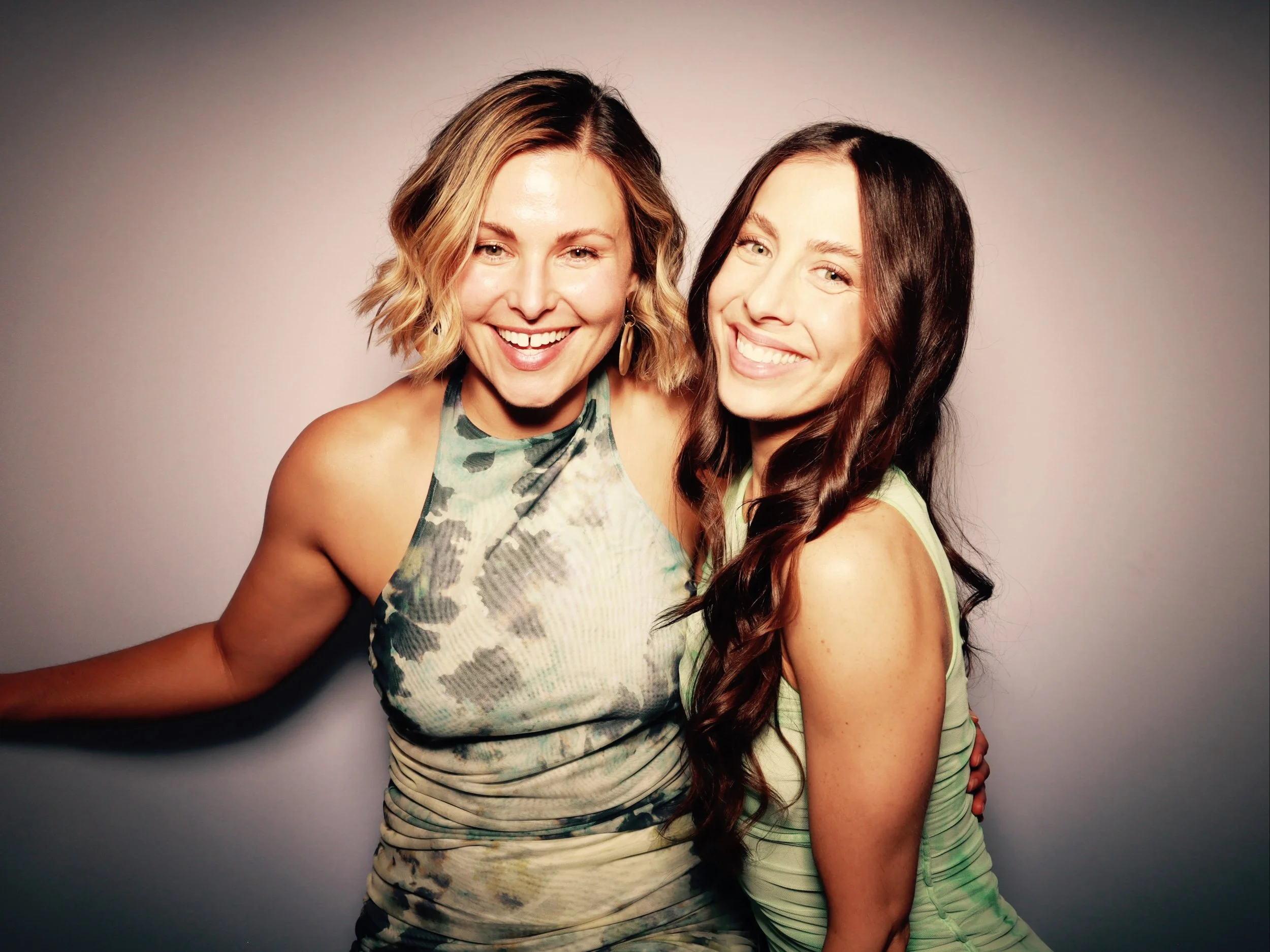 Two women smiling and hugging each other, one with short blonde hair and the other with long dark hair, against a plain background.