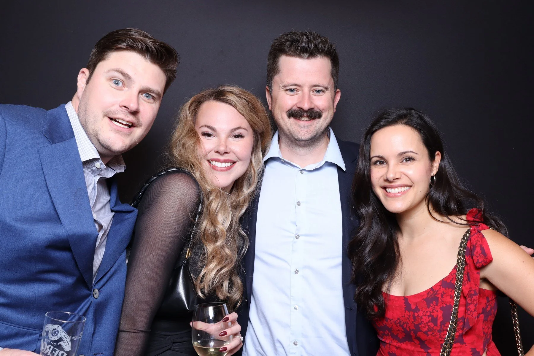 Group of four friends smiling and posing together at a celebration, one holding a glass of white wine, against a dark background.
