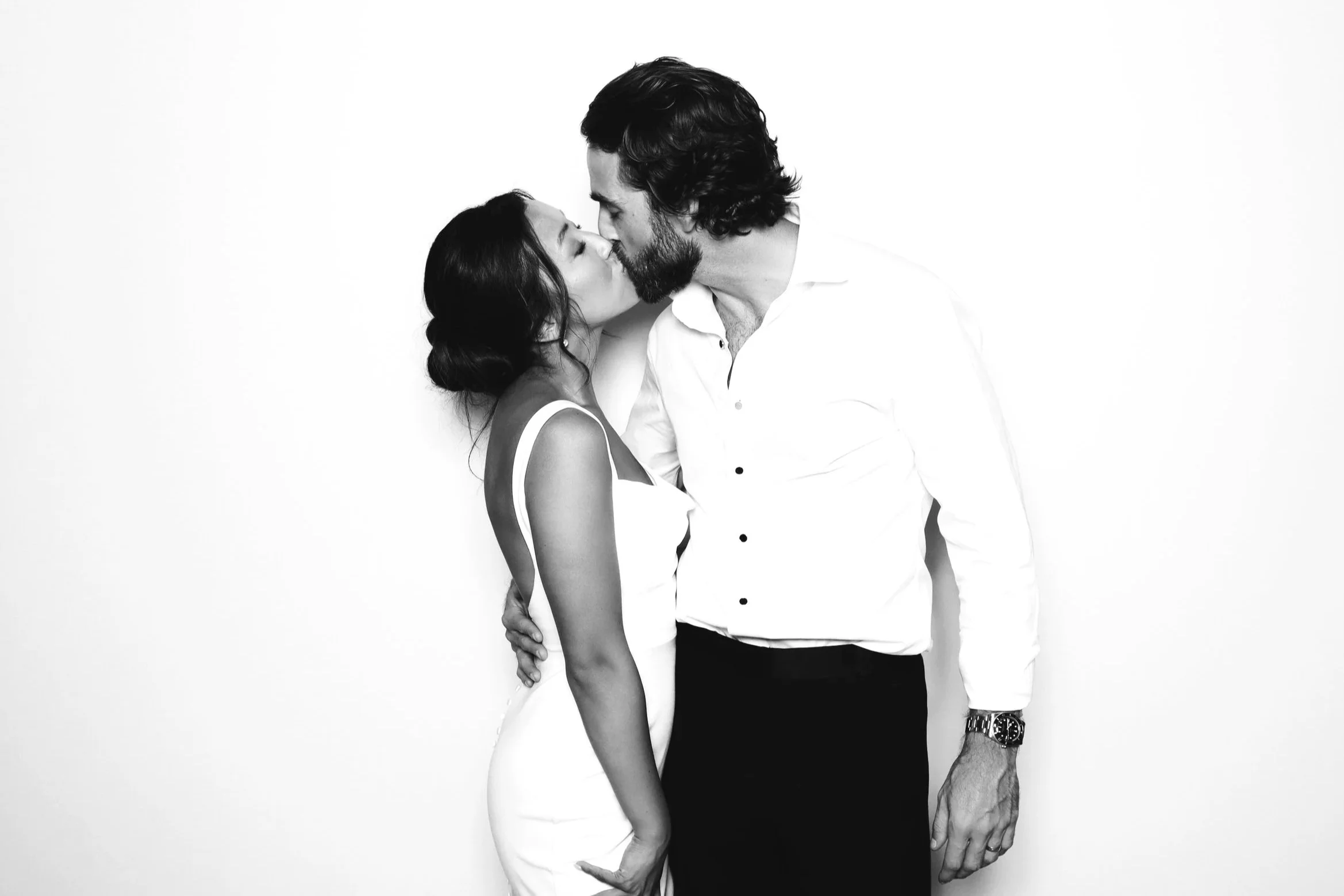 A black and white photo from a wedding photo booth rental featuring a bride in a white backless gown and a groom in a white button-down shirt kissing against a solid white backdrop.