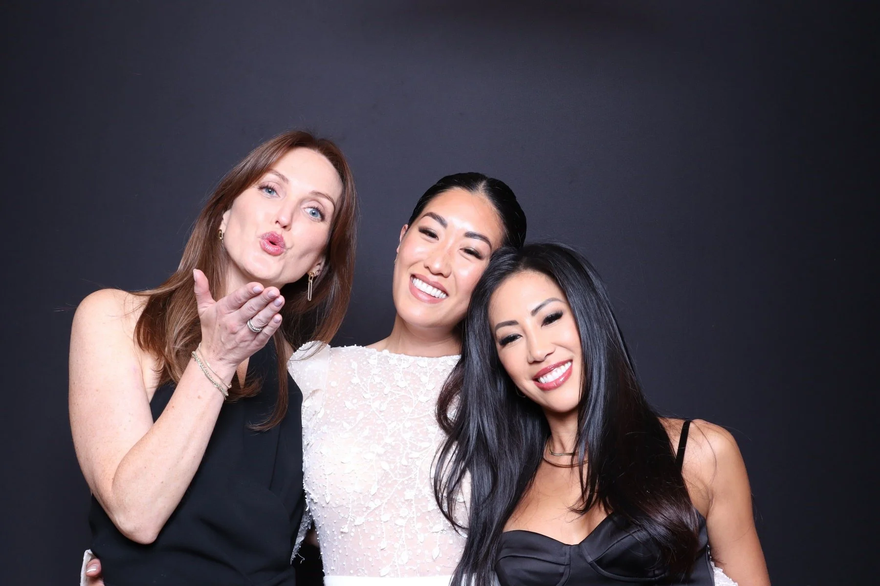 Three women smiling and posing together against a dark background, with the woman on the left blowing a kiss.