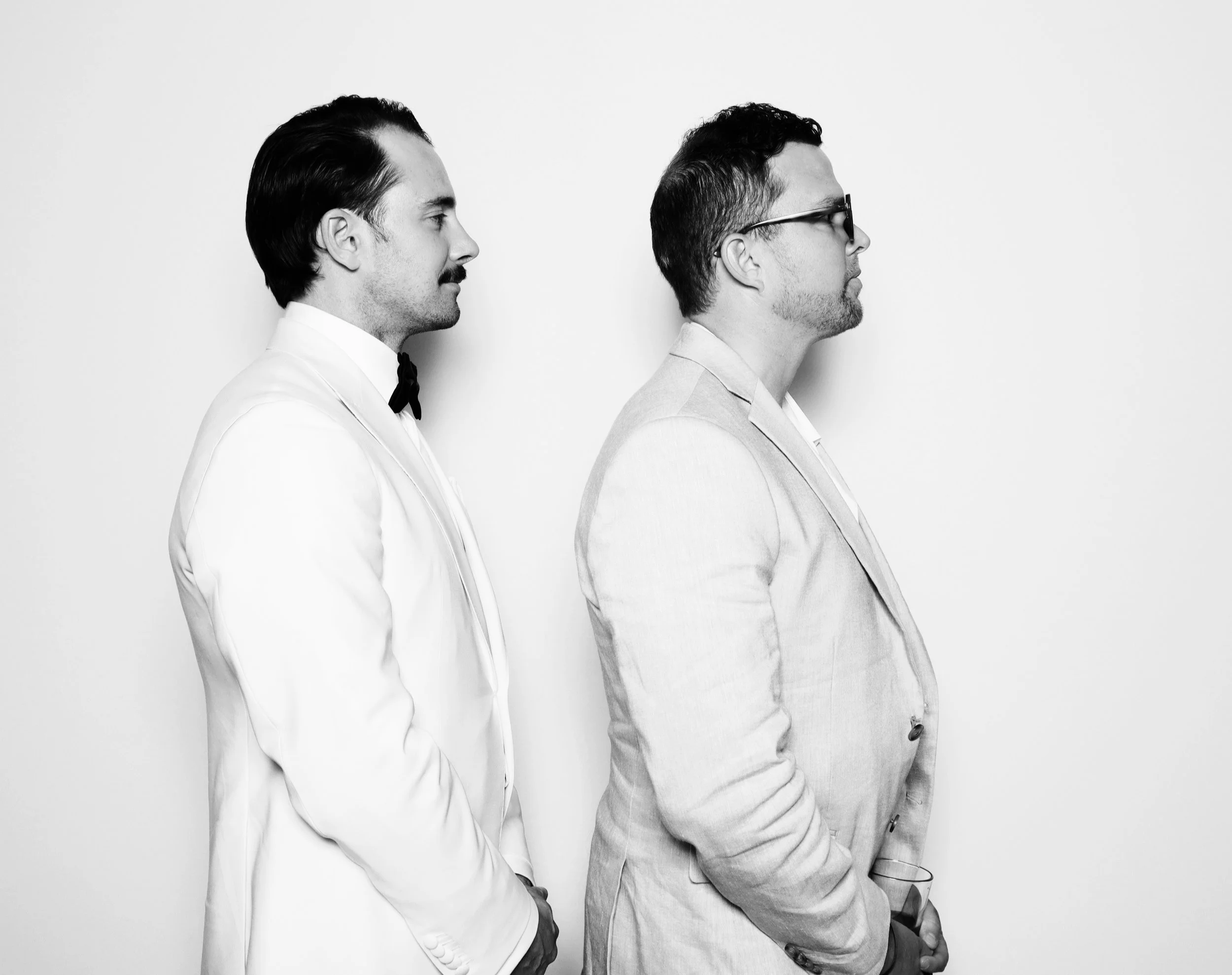 Two men in suits standing side by side against a plain white background, one with a bow tie and the other with glasses.