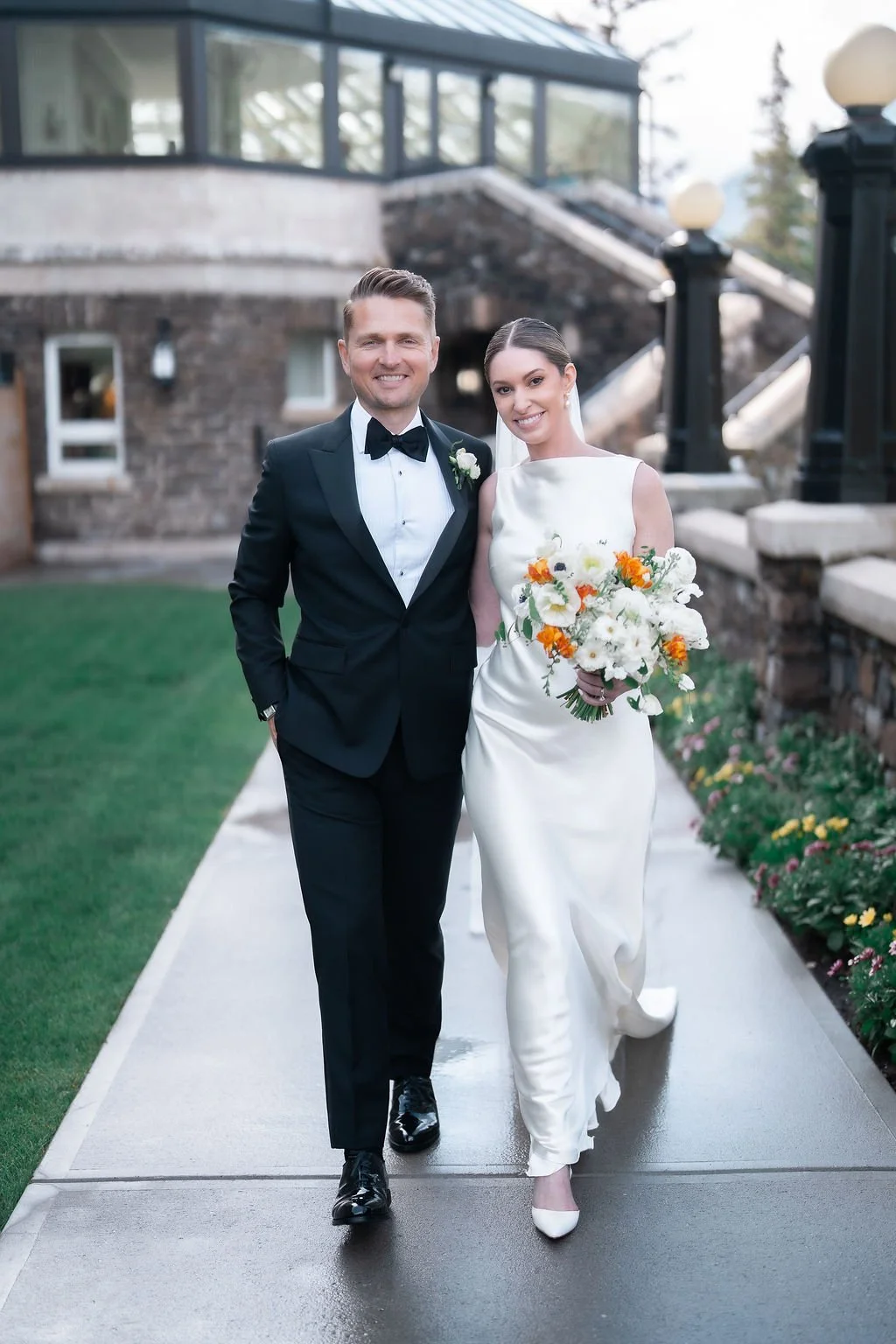 A newlywed couple walking outside on a cloudy day. The groom is wearing a black tuxedo with a bow tie, and the bride is in a white satin wedding gown holding a bouquet of white, orange, and yellow flowers. They are smiling and walking on a sidewalk w