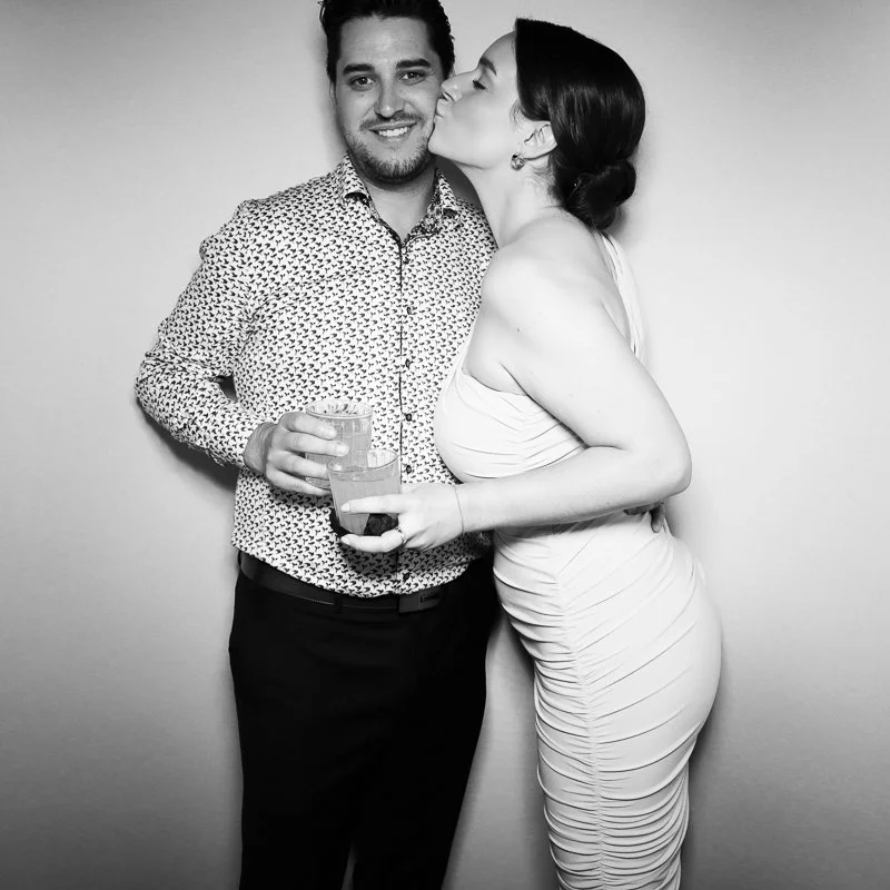 A man and woman at a celebration; the woman kisses the man's cheek while the man smiles, holding two drinks in his hand.