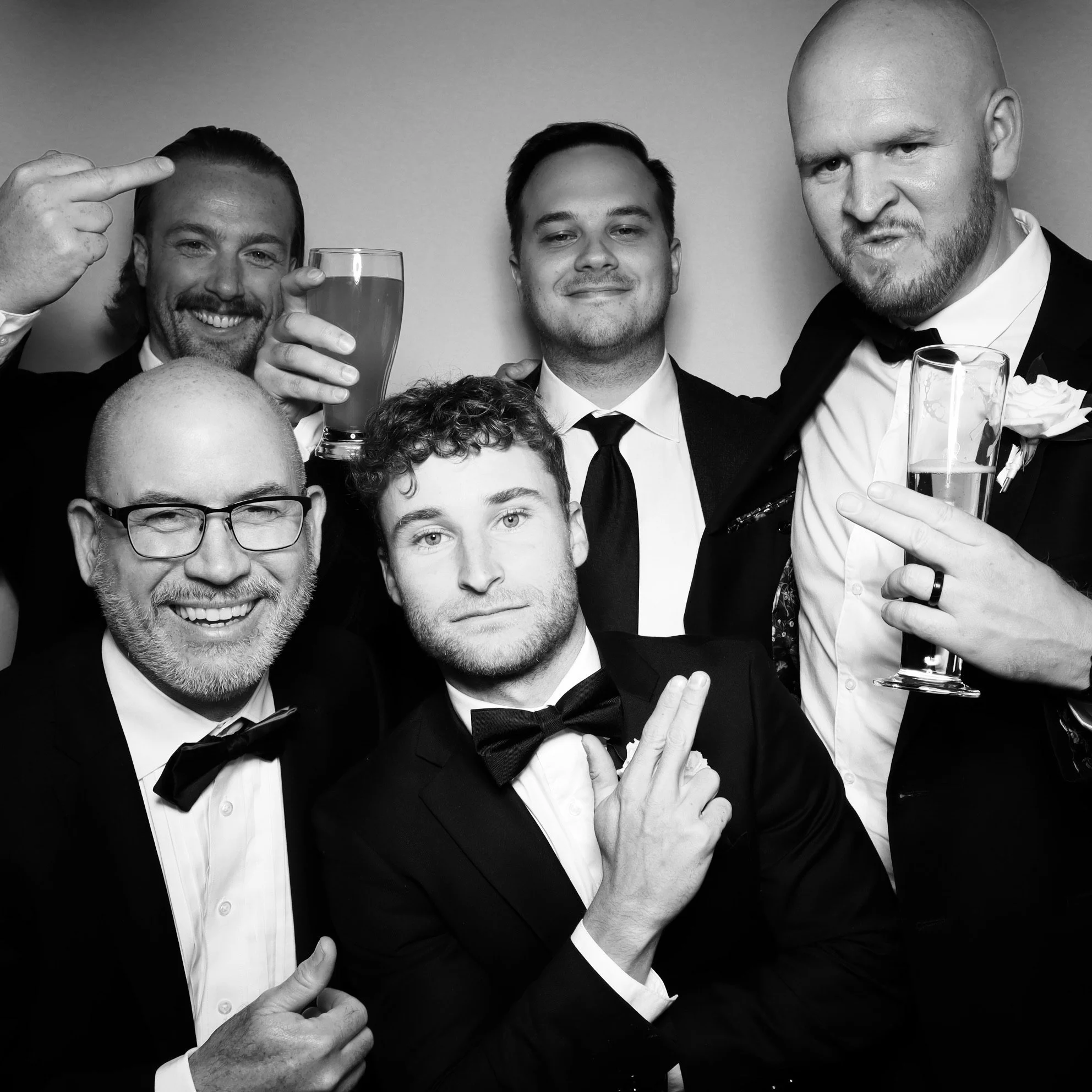 Group of six men dressed in tuxedos at a formal event, some holding drinks, smiling and posing for a photo in black and white.