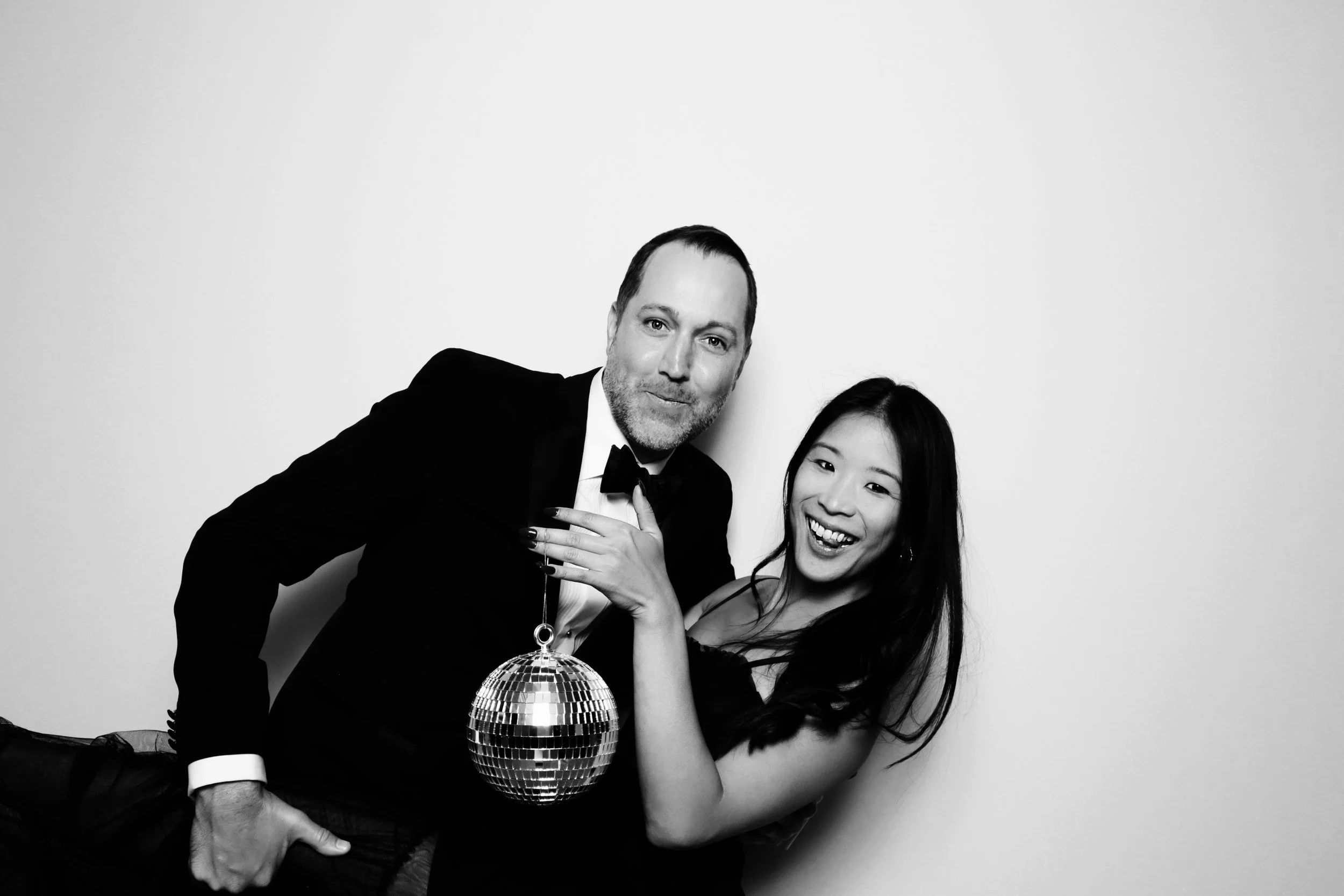 A man and a woman in formal attire posing with a disco ball against a plain white background, both smiling and looking at the camera.