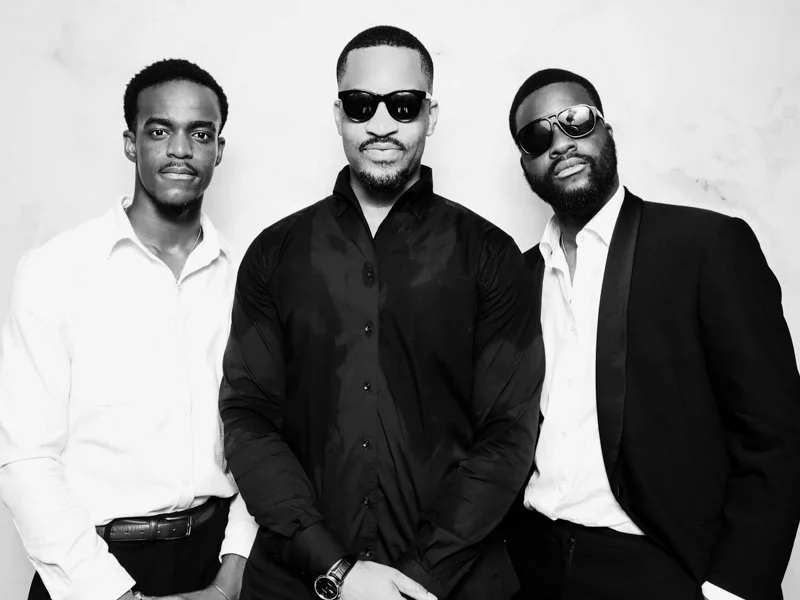 Three men standing together, dressed in stylish clothing, with two wearing sunglasses, in front of a plain background.
