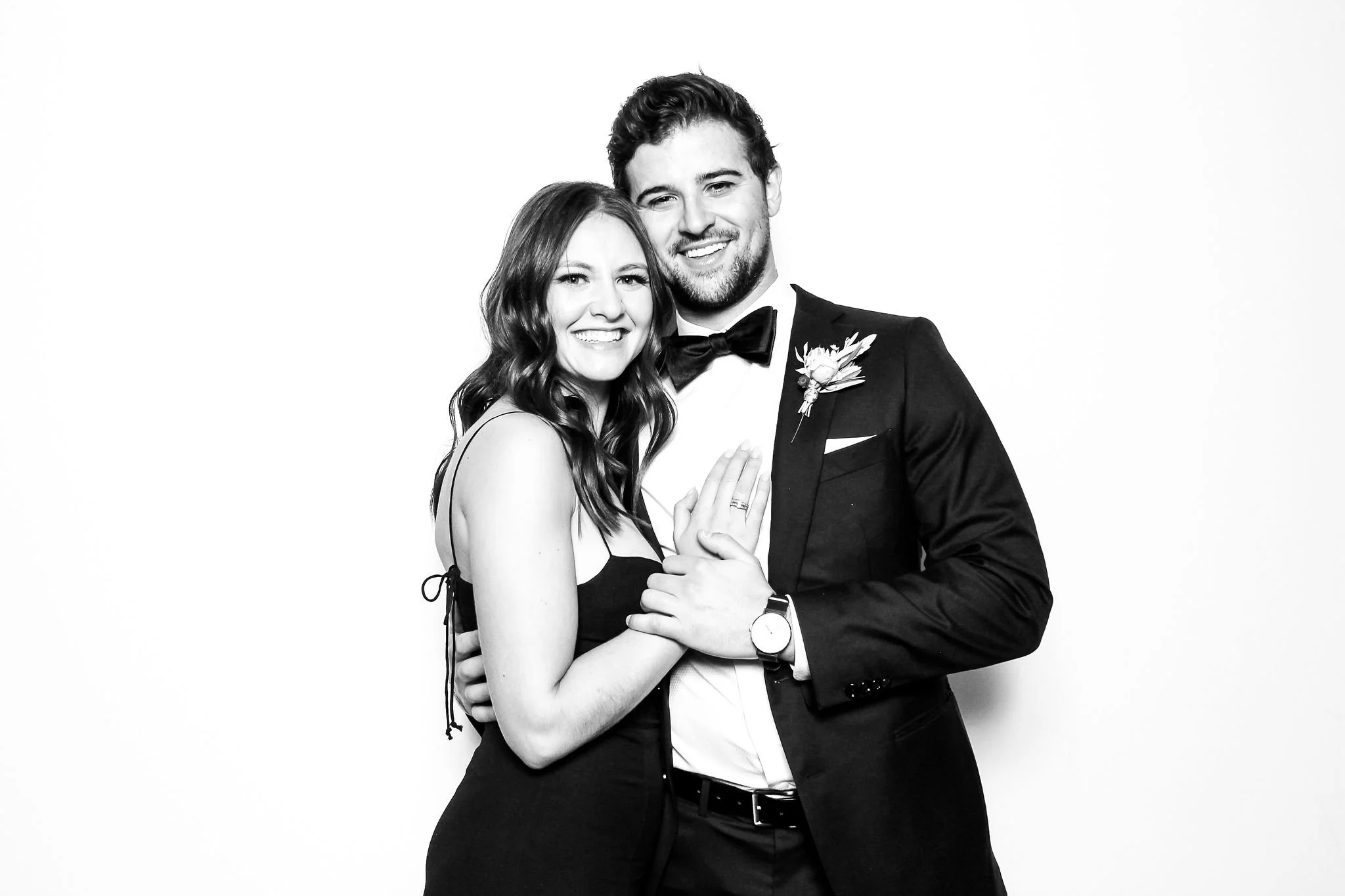 A smiling couple dressed in formal attire, the woman in a black dress and the man in a tuxedo, standing close together against a plain white background.