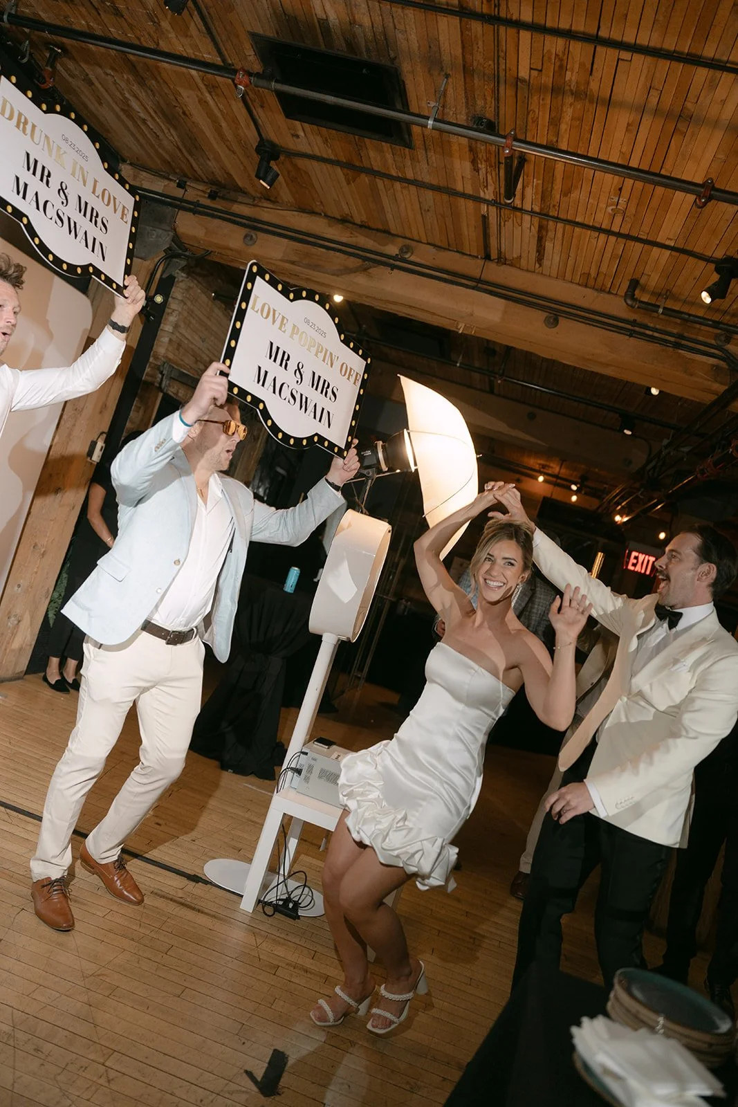 A bride and groom dancing together at their wedding reception, surrounded by guests holding signs that read 'LOVE POPPIN' OFF' and 'DRUNK IN LOVE MR & MRS MACSWAIN'. The bride is smiling and wearing a strapless white dress, while the groom is wearing