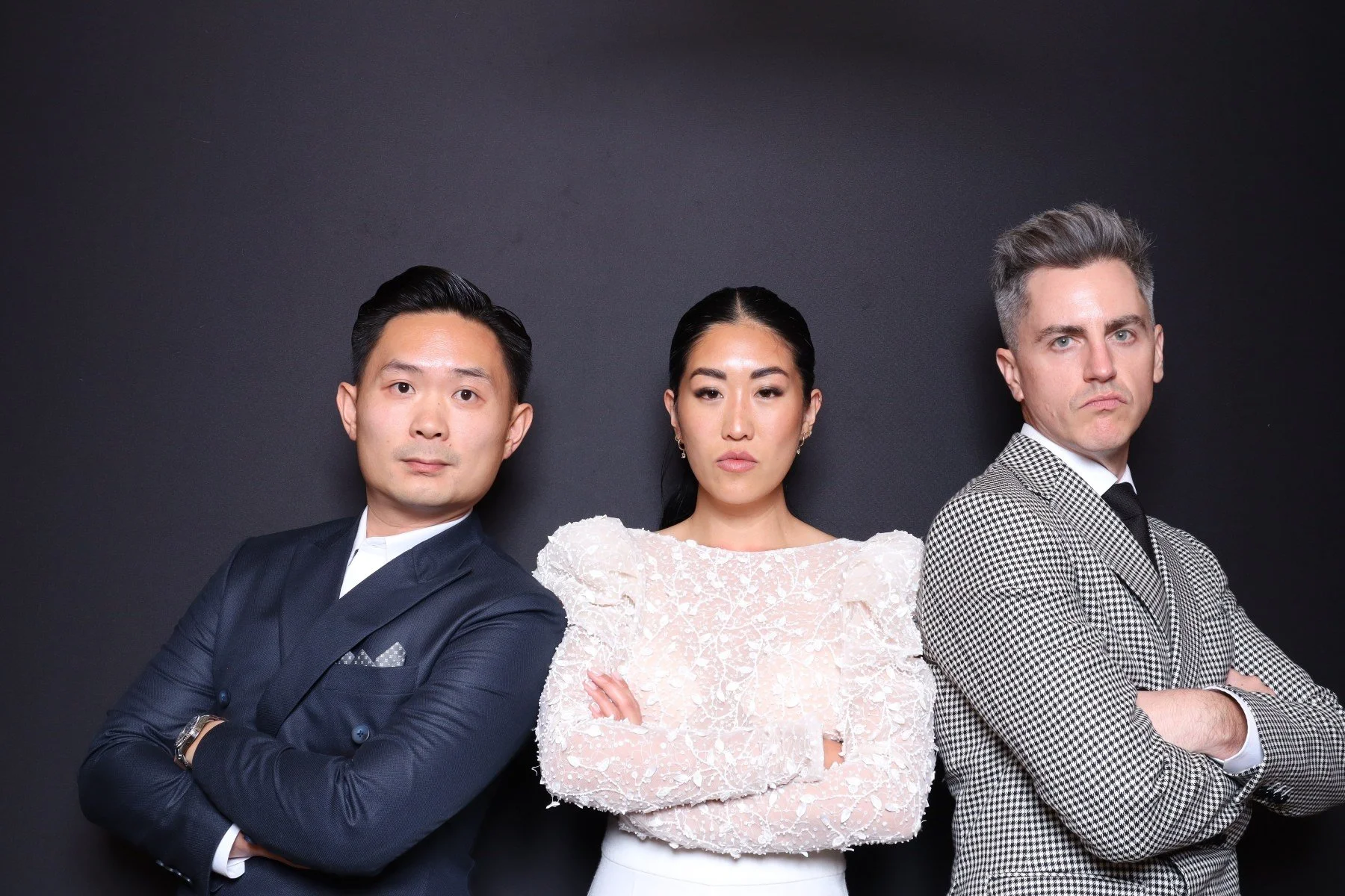 Three professionally dressed individuals, two men and one woman, standing against a dark background with arms crossed, looking serious.