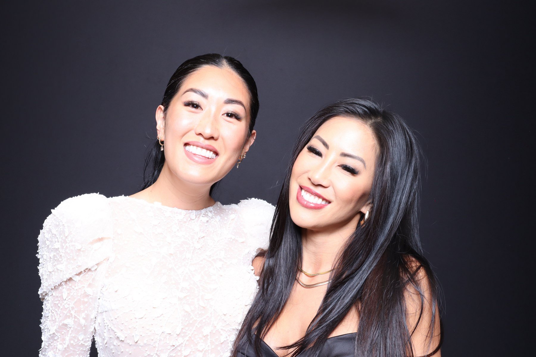 Two women smiling together against a dark background, one wearing a white textured dress with puffed sleeves and the other wearing a black strapless top with layered necklaces.