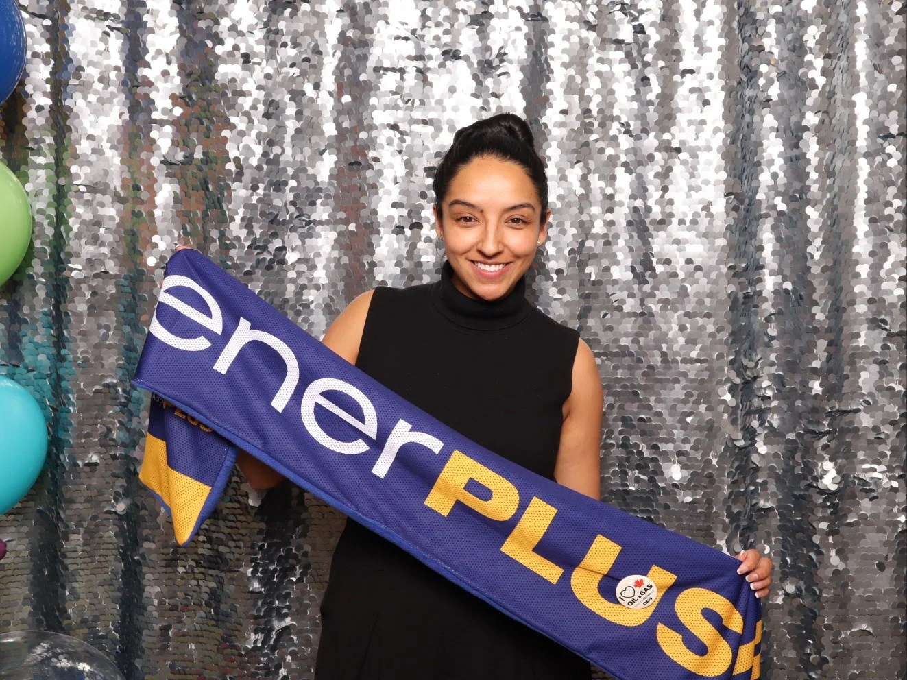 A woman smiling and holding a blue and yellow scarf that reads 'Premier Plus' in front of a shiny silver sequin backdrop with balloons on the side.