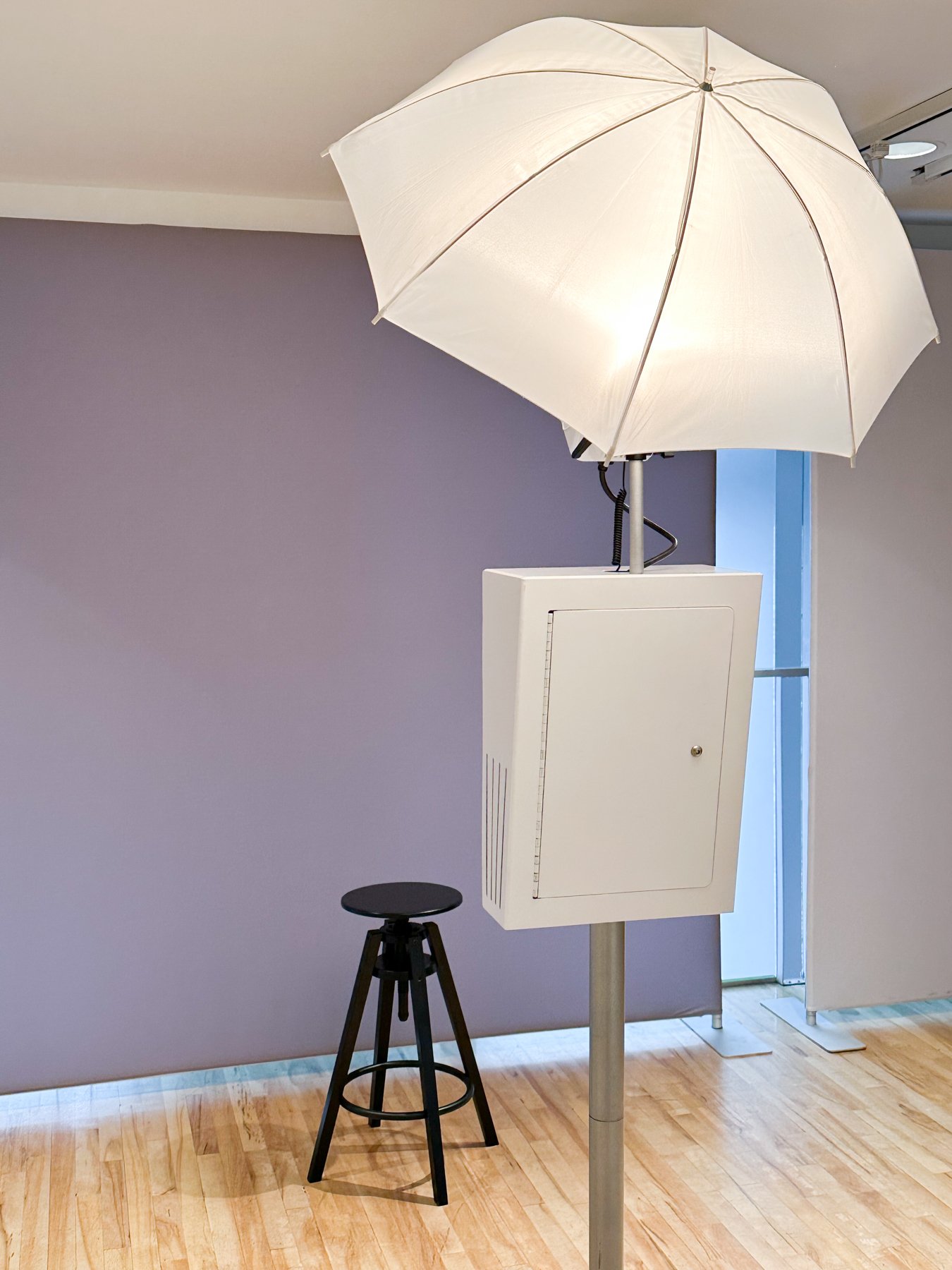 Studio photography setup with a large white umbrella light, a white electrical box on a stand, and a black stool on a hardwood floor.