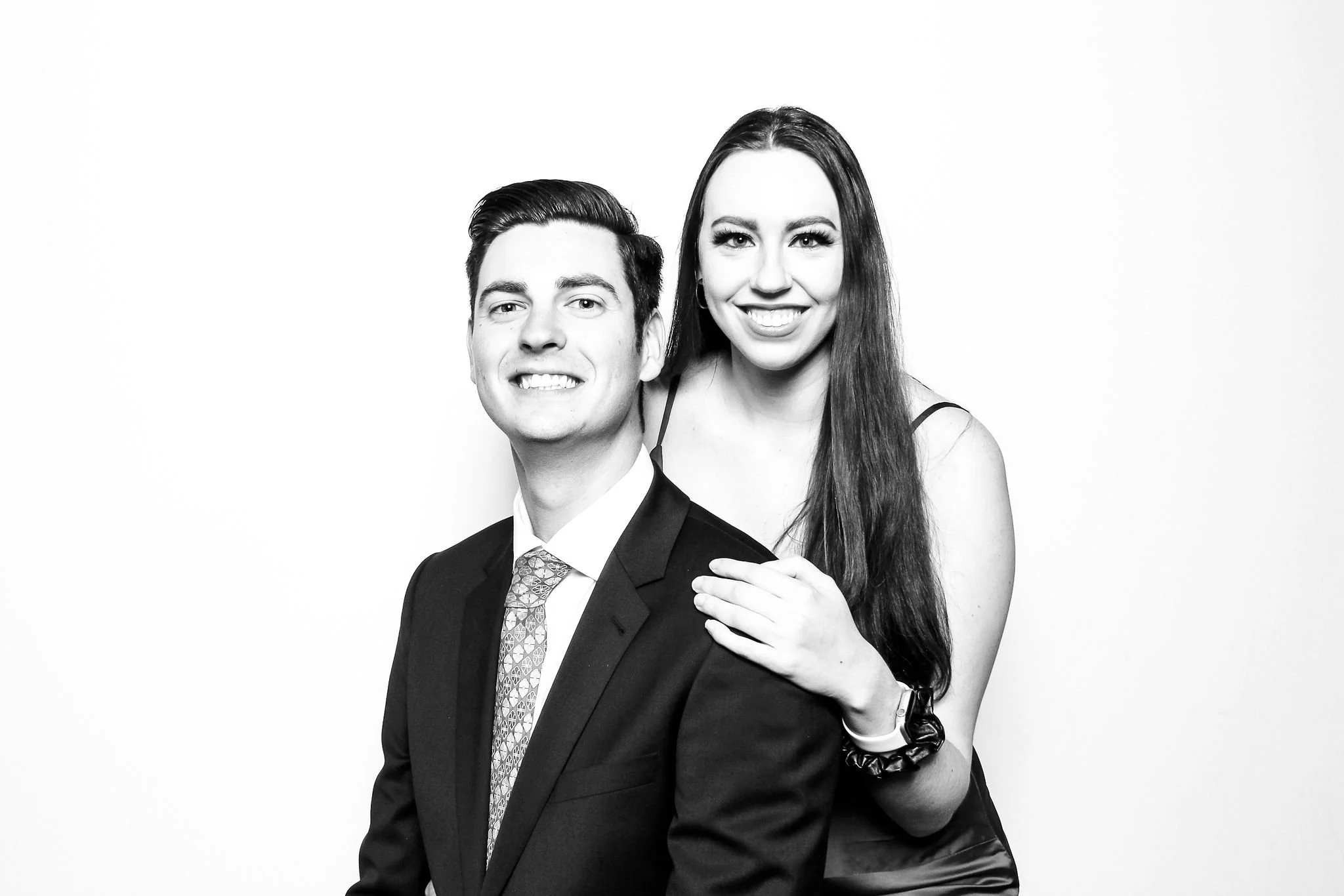 A black and white photo of a smiling man in a suit and a woman with long hair, wearing a sleeveless top, standing close together against a plain light background.