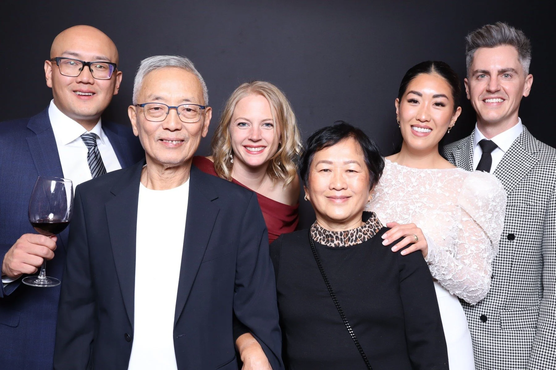Group of six diverse people at a formal gathering, smiling at the camera. One person is holding a glass of red wine. They are dressed in business or party attire, standing against a dark background.