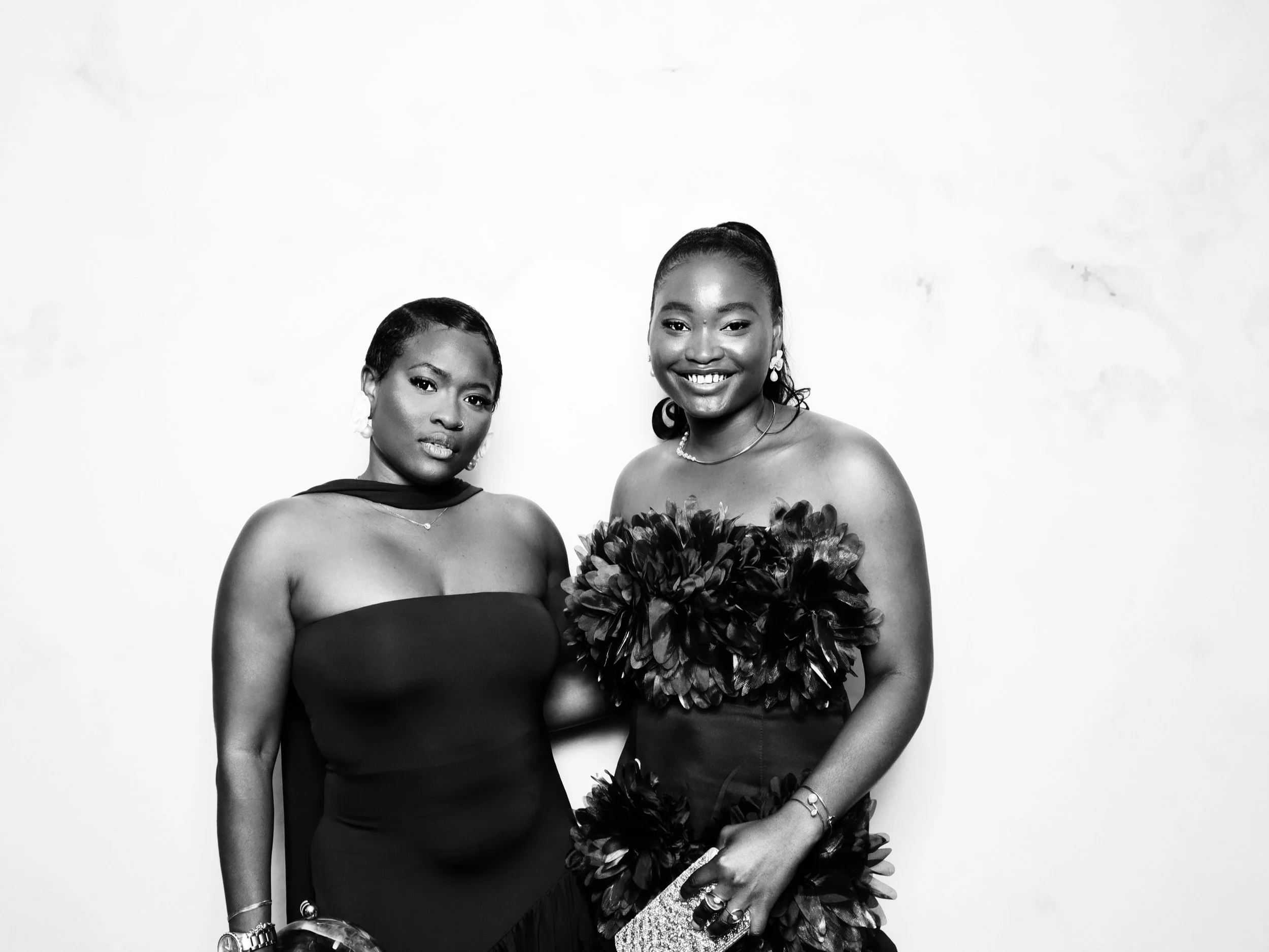 Two women dressed in elegant evening gowns standing against a plain backdrop, smiling at the camera.