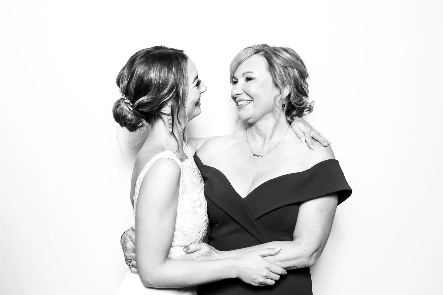 A woman and a young girl are smiling and looking at each other, with their arms around each other, against a plain background.