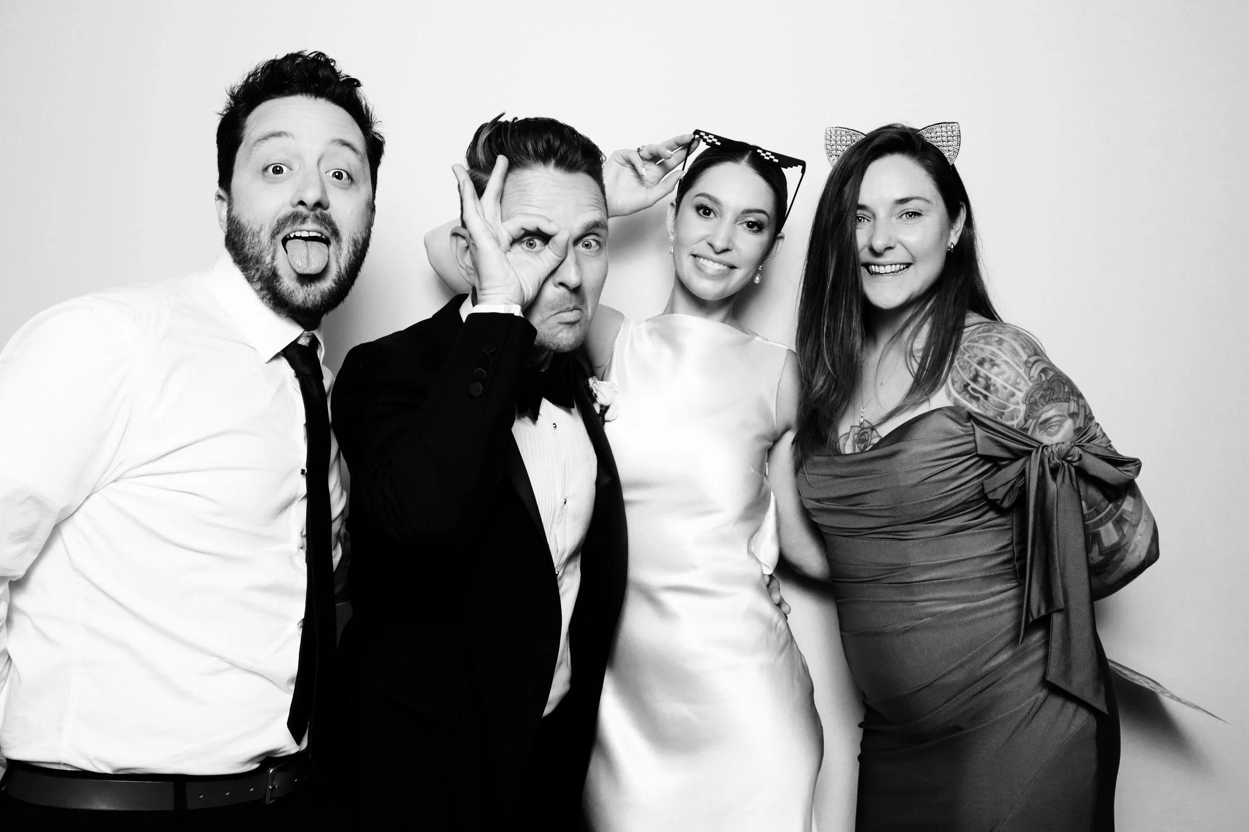Four people at a formal event, smiling and posing against a plain backdrop, with one person making a gesture with their hand.